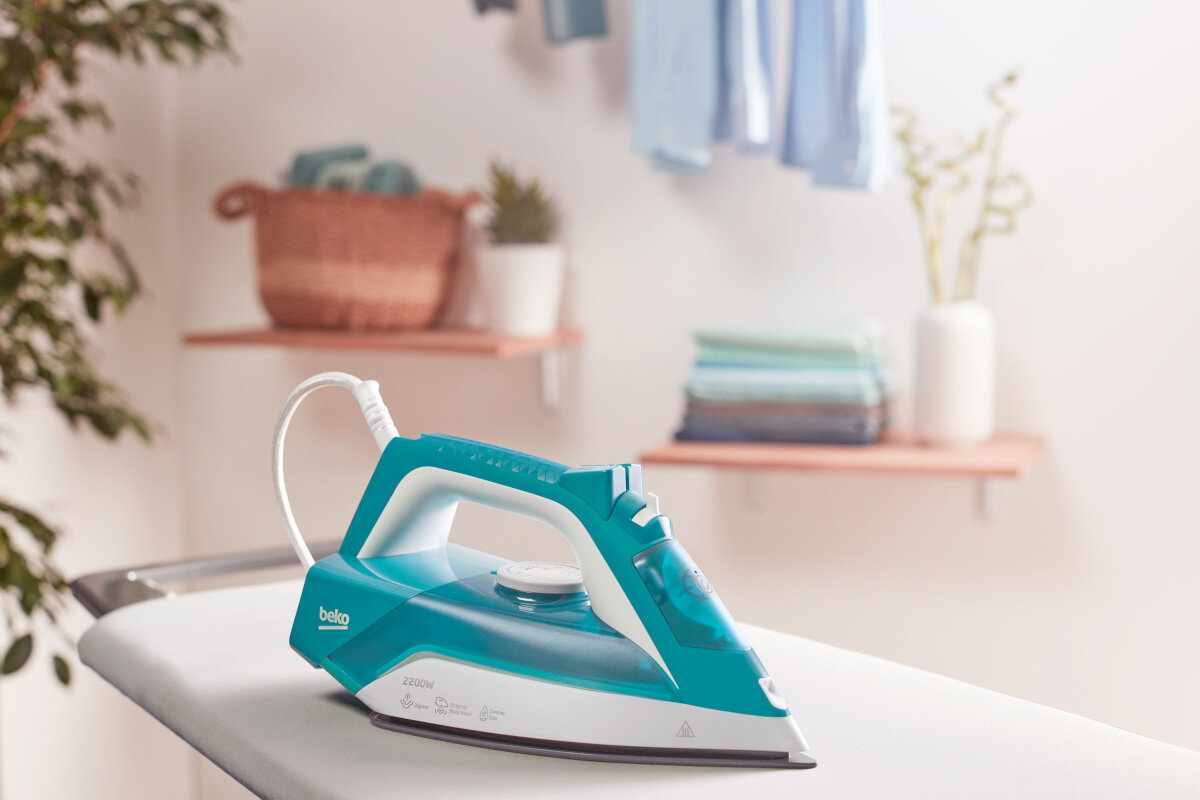 Turkusowe i białe żelazko na desce do prasowania. Pranie i półki w tle.