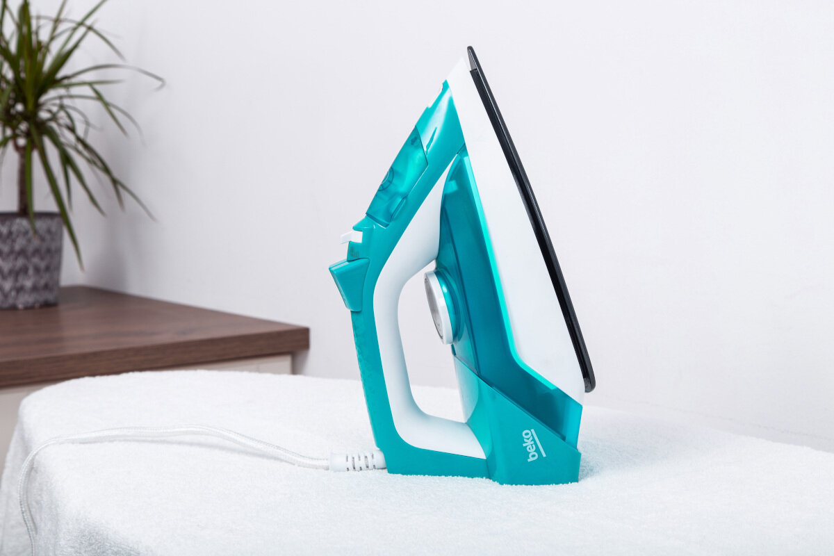 Turkusowe żelazko na białym ręczniku, białe tło z rośliną, widok z boku.