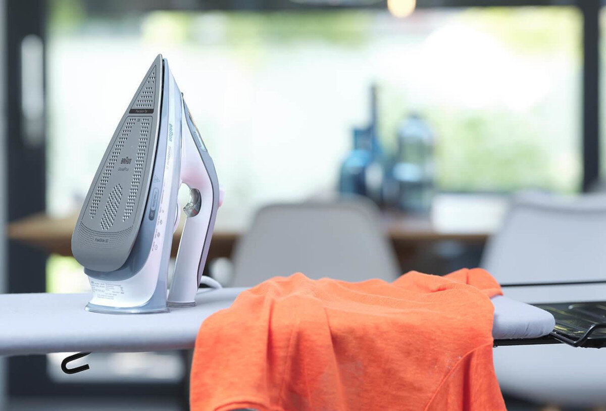 Żelazko na desce do prasowania z pomarańczową koszulą. Żelazko jest białe i szare.