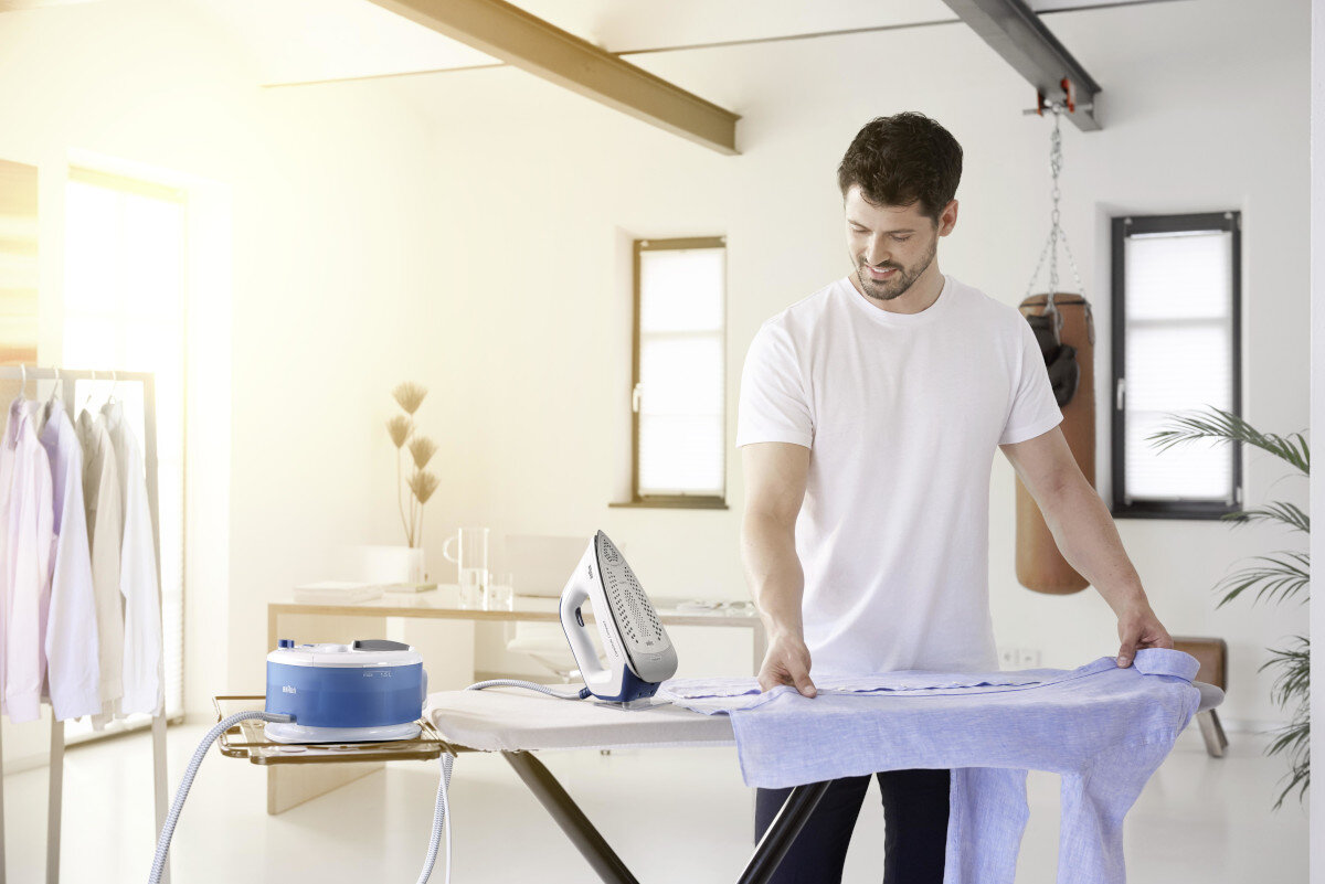 Mężczyzna prasujący koszulę na desce do prasowania z żelazkiem parowym i wieszakiem.