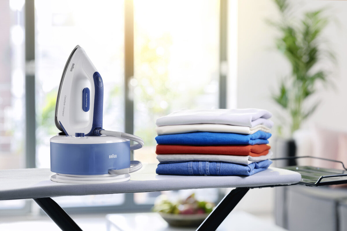 Ustawienie do prasowania z białym i niebieskim żelazkiem na niebieskiej podstawie, ubraniami i deską do prasowania, z oknem z tyłu.