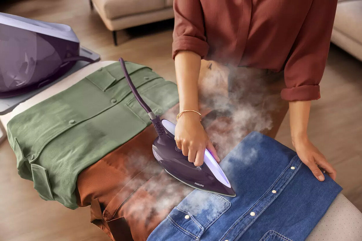 Osoba prasuje dżinsowe koszule parą. Inne koszule są na desce do prasowania. Fioletowa parownica do ubrań.