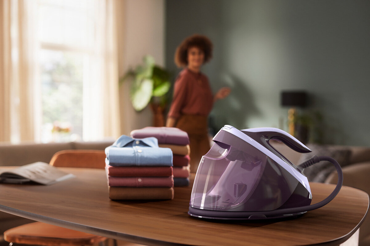 Kobieta jest w tle, stos złożonych ubrań i fioletowa parownica do ubrań na stole.