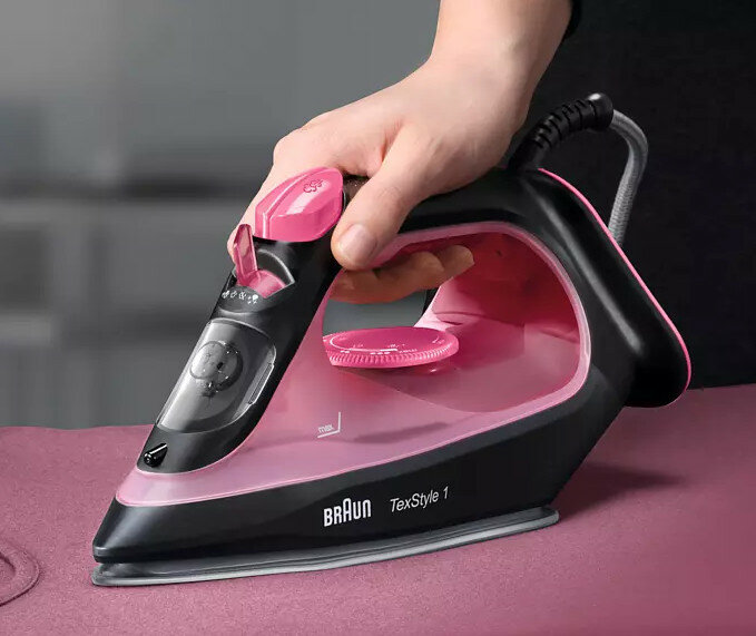 Czarne i różowe żelazko. Dłoń trzymająca żelazko nad różową tkaniną. Widoczne elementy sterujące i logo Braun.