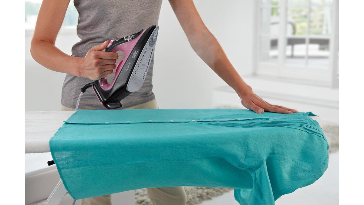 Osoba prasująca turkusową koszulę na białej desce do prasowania, w tle okno.