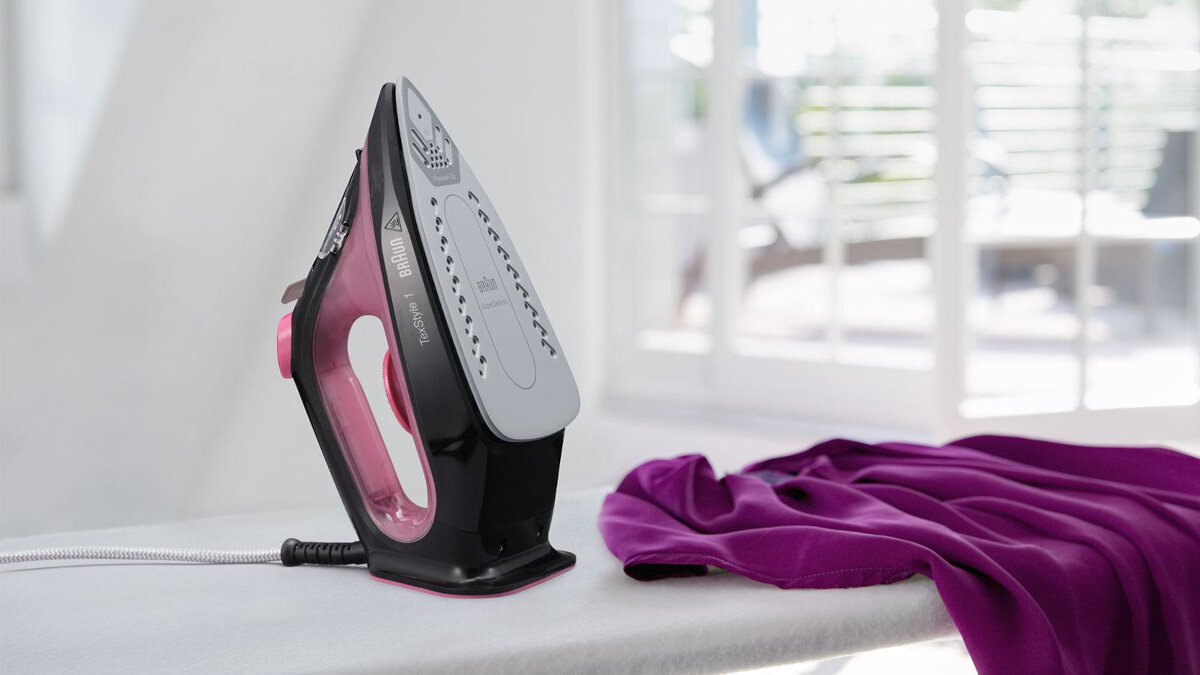 Czarne i różowe żelazko na desce do prasowania. Fioletowa tkanina po prawej. Biała ściana i okno.