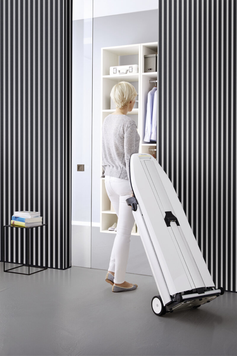 Kobieta tocząca białą deskę do prasowania na kółkach. Pionowa ściana i podłoga w paski.