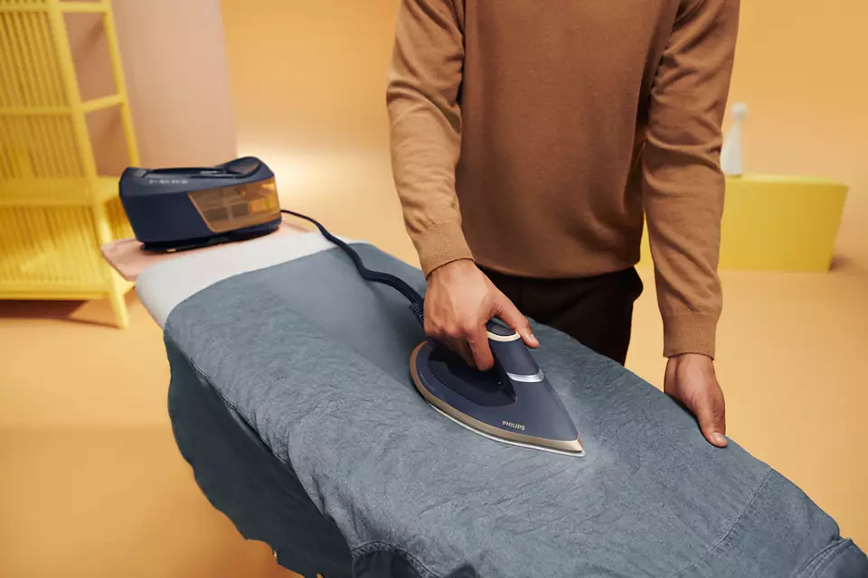 Mężczyzna prasuje szarą koszulę na desce do prasowania. Żelazko jest ciemnoniebieskie i złote.