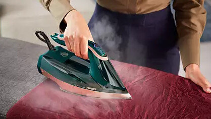 Kobieta prasuje czerwoną tkaninę zielono-różowym żelazkiem, para unosi się. Tło rozmyte.