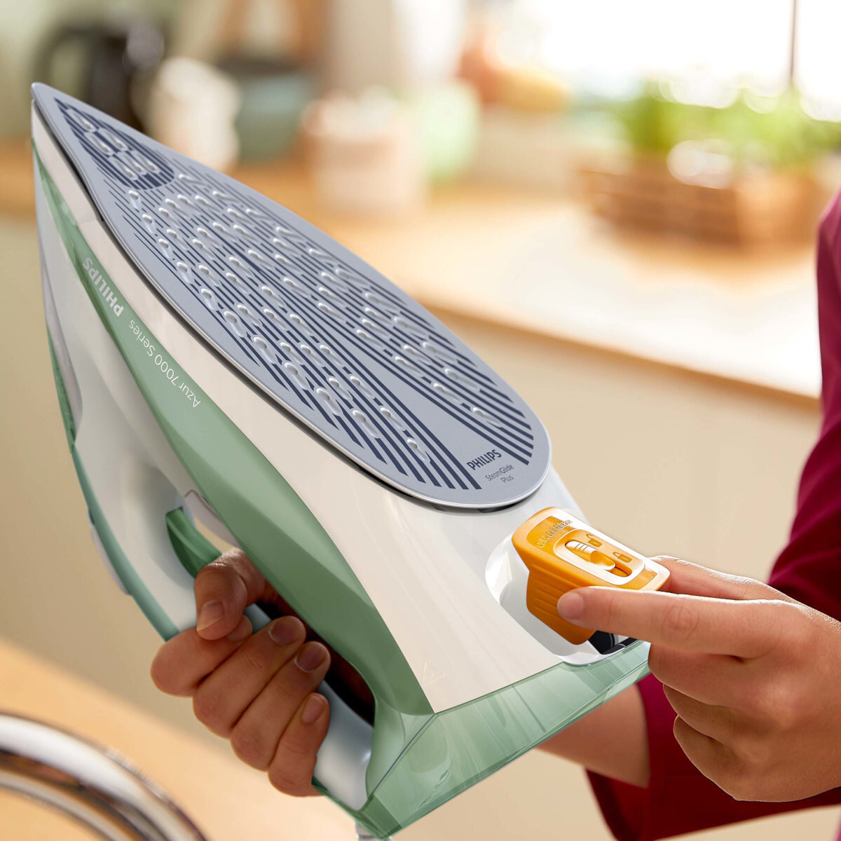 Ręka osoby obsługująca zielono-białe żelazko z pomarańczowym pokrętłem.