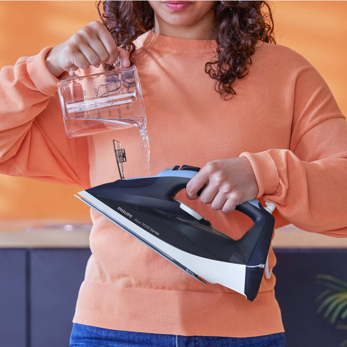 Osoba wlewa wodę do żelazka. Żelazko jest czarne i niebieskie. Pomarańczowe tło swetra.