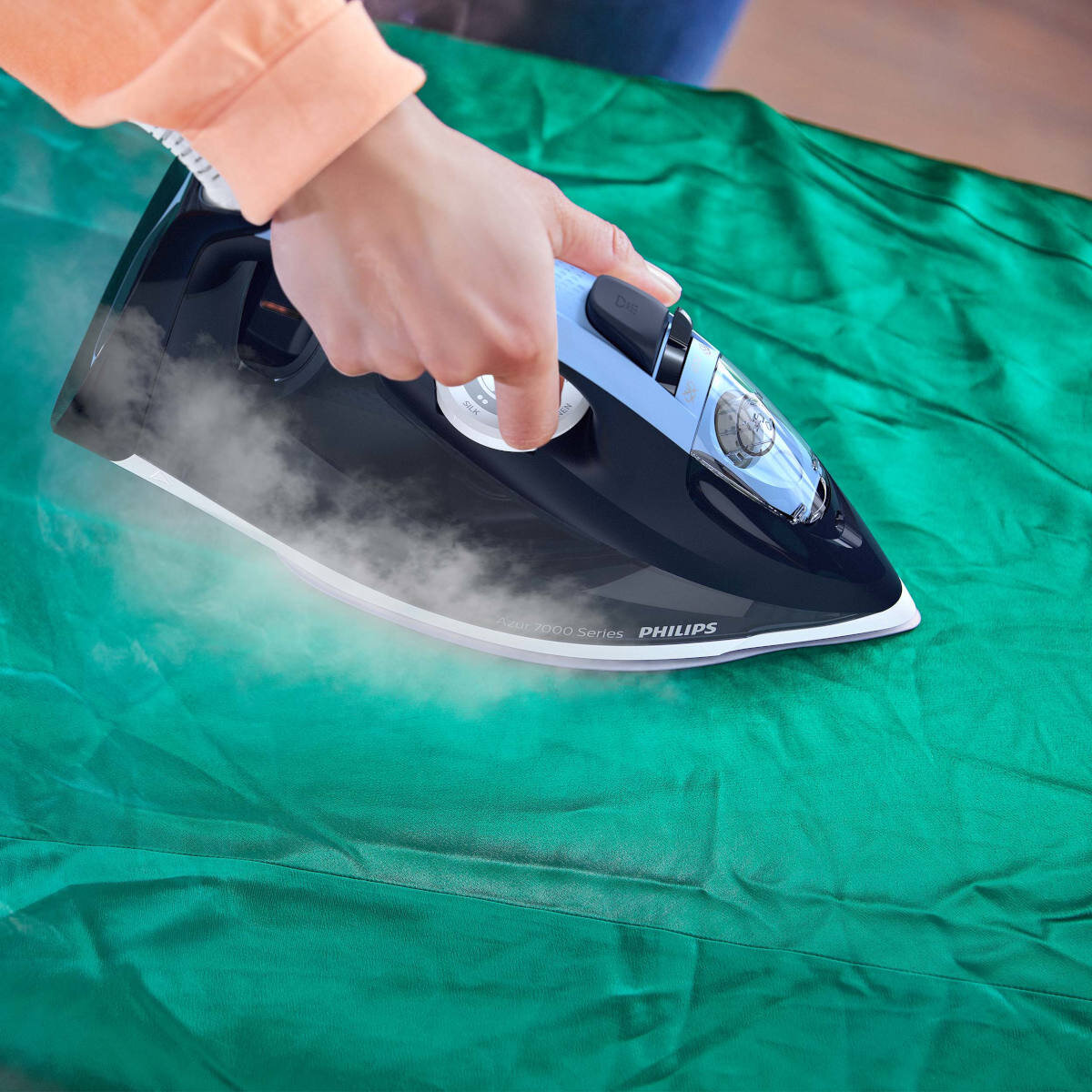 Osoba prasująca zielony materiał niebiesko-czarnym żelazkiem, widoczna para.