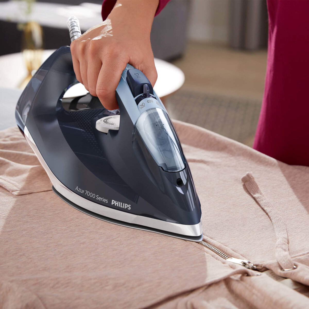 Osoba prasuje różową kurtkę. Czarno-niebieskie żelazko, białe tło.