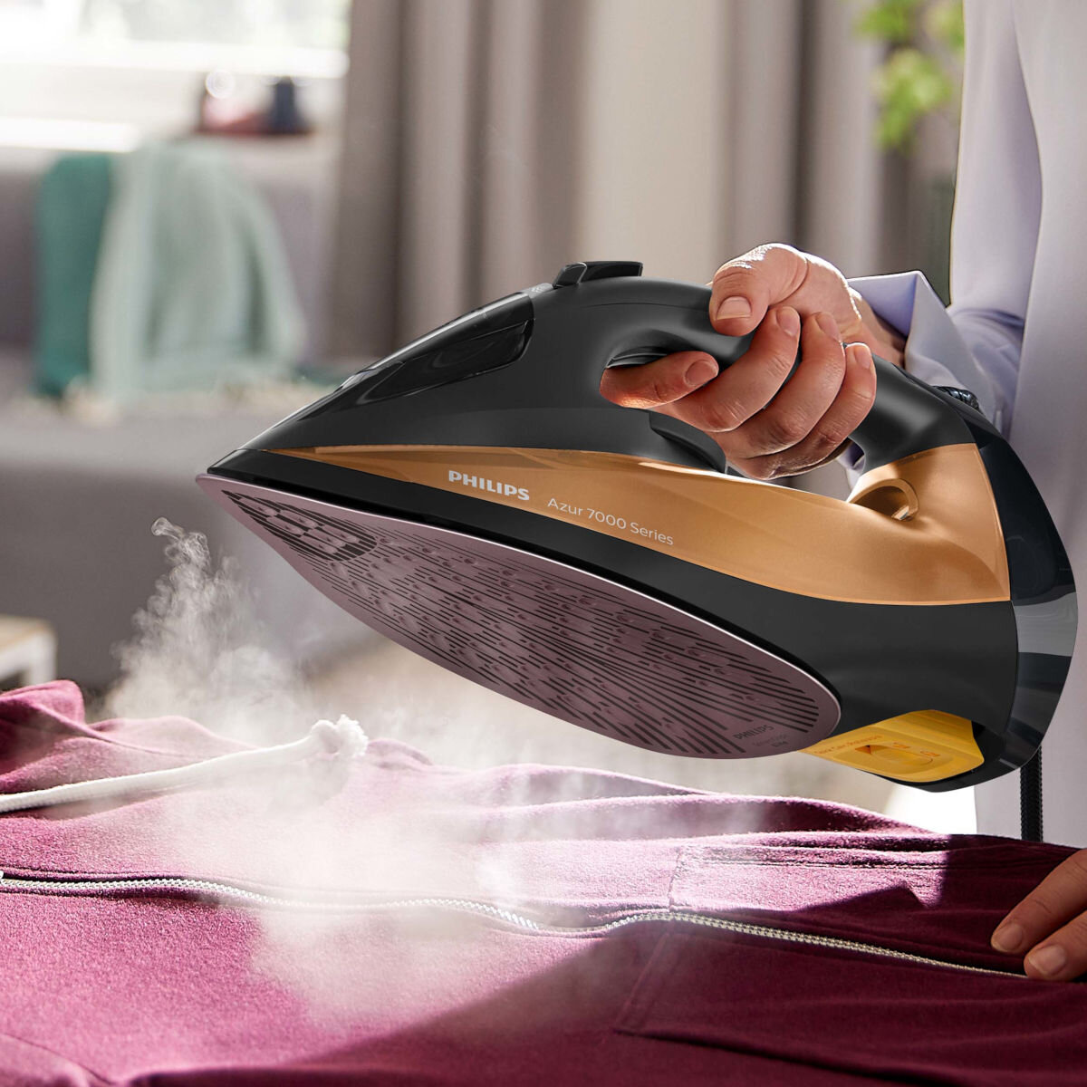 Osoba prasująca bordową bluzę z kapturem czarno-złotym żelazkiem, para unosi się.