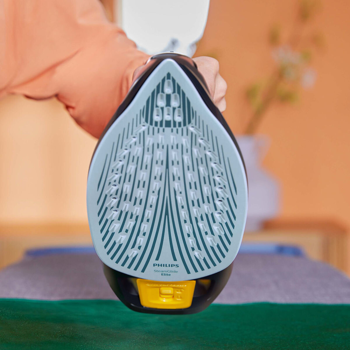 Zbliżenie płyty żelazka, widok z przodu. Dłoń trzymająca żelazko. Turkusowy i czarny design.