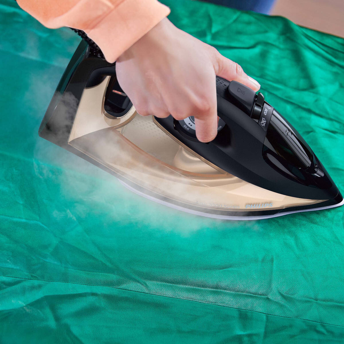 Ręka prasująca zieloną tkaninę czarnym żelazkiem, wytwarzająca parę, pokazująca markę Phillips.