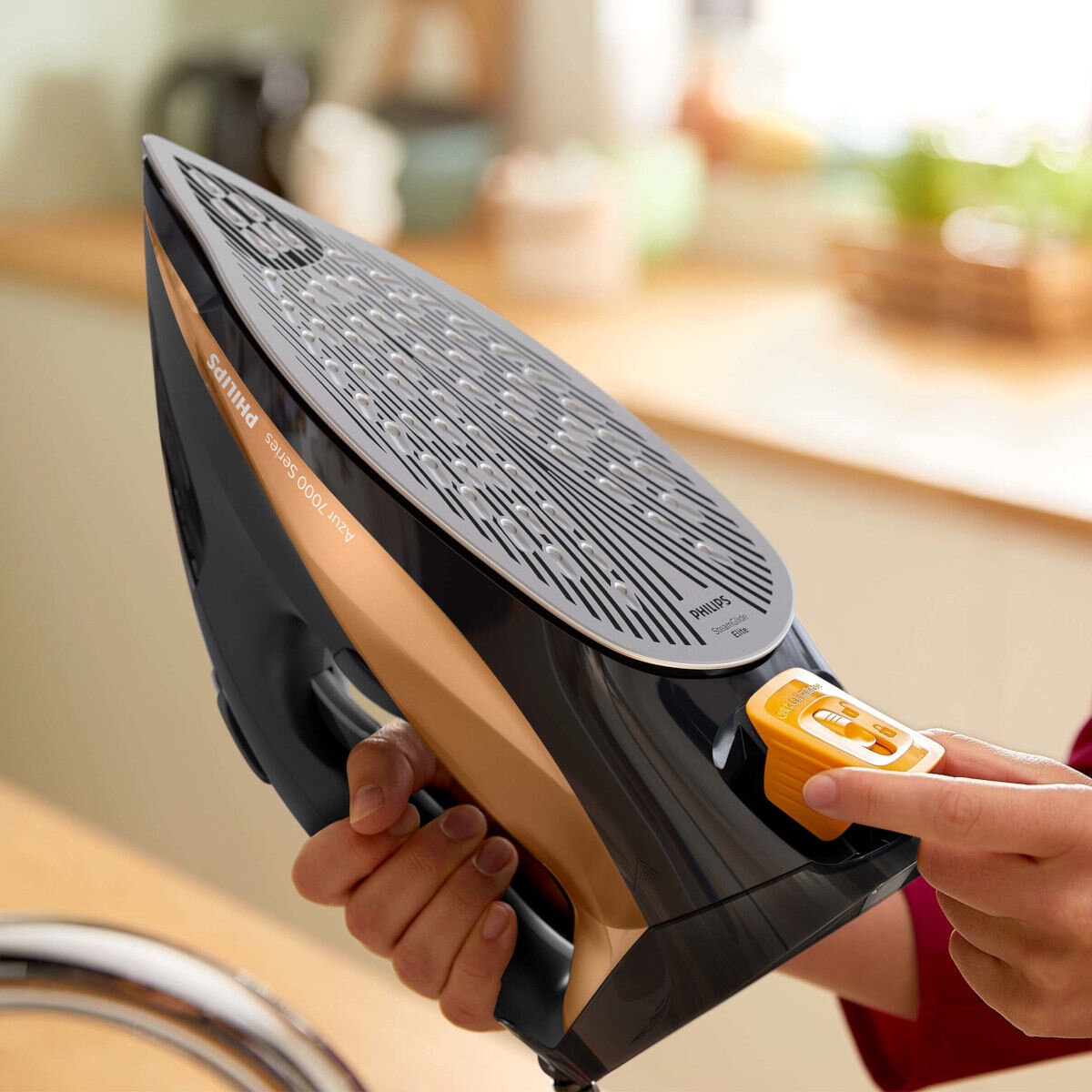 Czarne i złote żelazko. Ręka trzyma żelazko. Żelazko znajduje się na białej powierzchni.