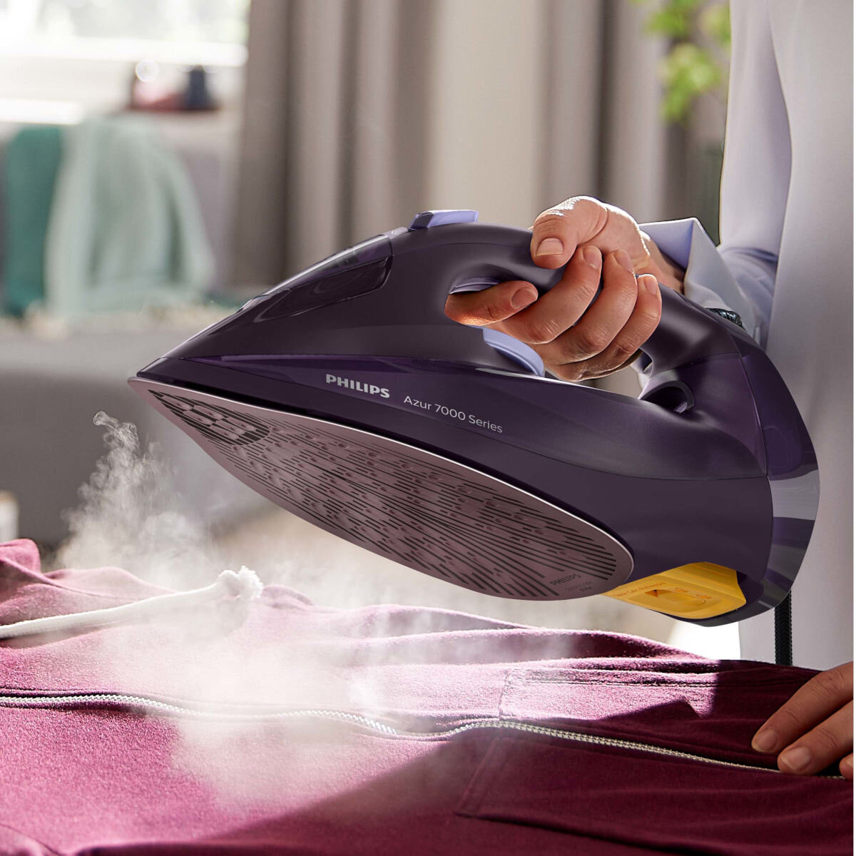 Osoba prasuje bordową bluzę z kapturem fioletowym żelazkiem. Para jest widoczna.
