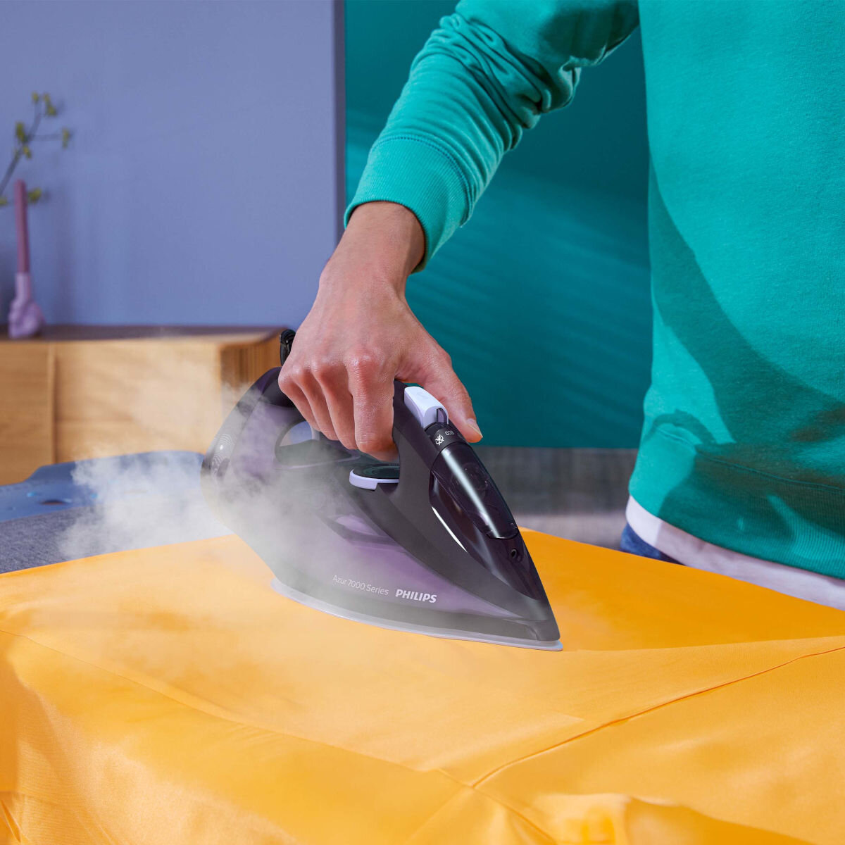 Osoba prasująca żółtą tkaninę czarnym i fioletowym żelazkiem, widoczna para.