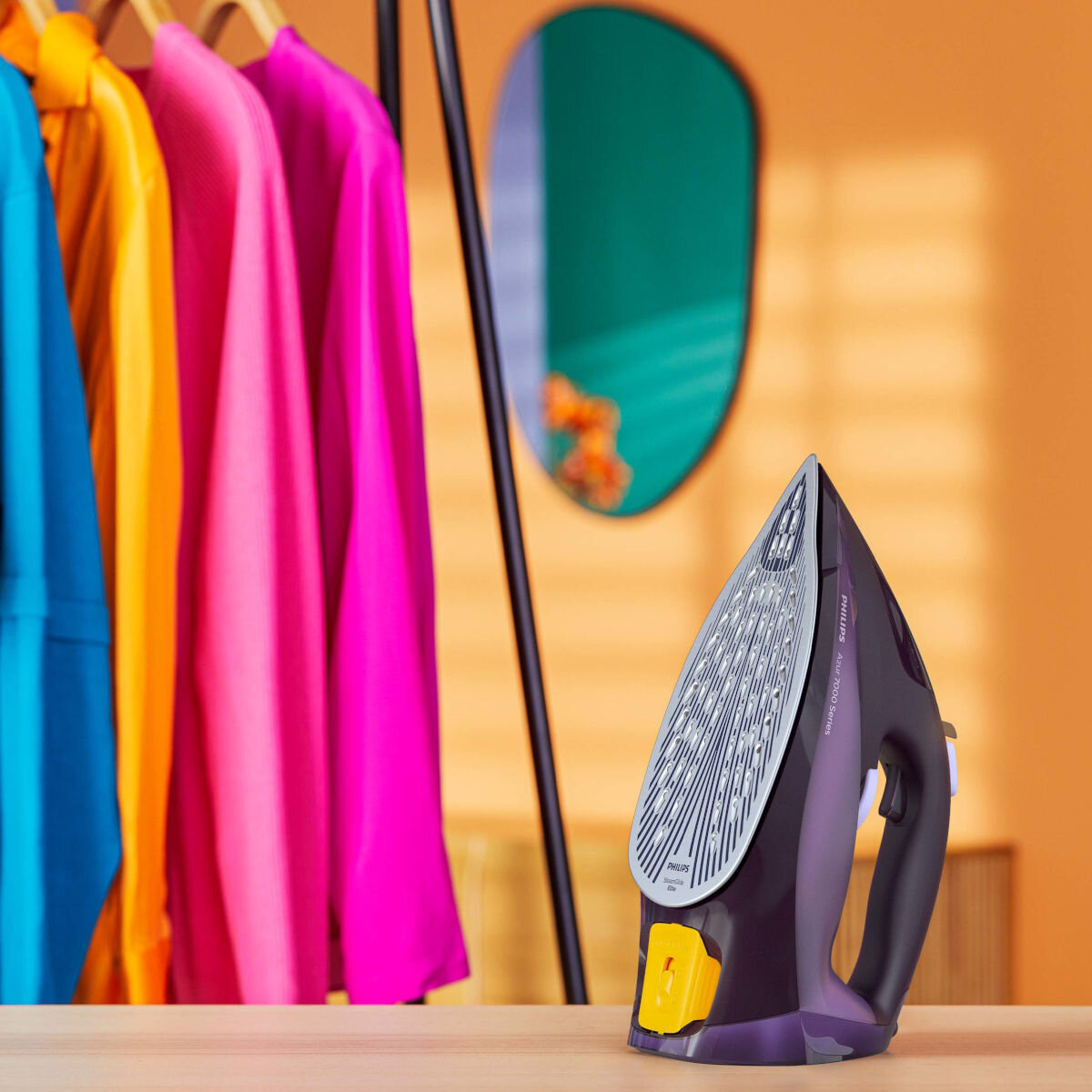 Żelazko i wieszak na ubrania z kolorowymi ubraniami. Pomarańczowa ściana i zielone lustro w tle.