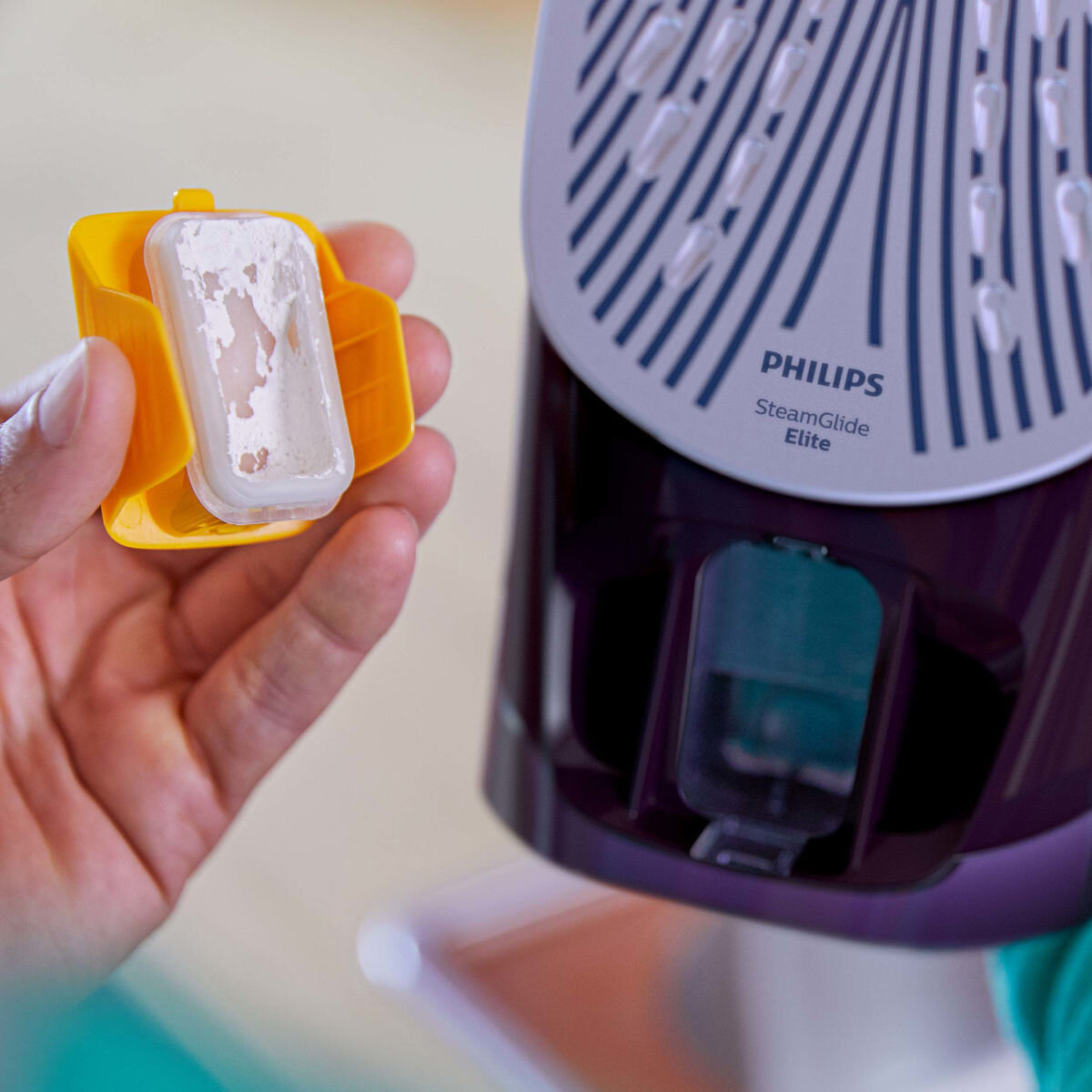 Dłoń trzymająca żółty pojemnik z zużytym wkładem odkamieniającym obok żelazka.