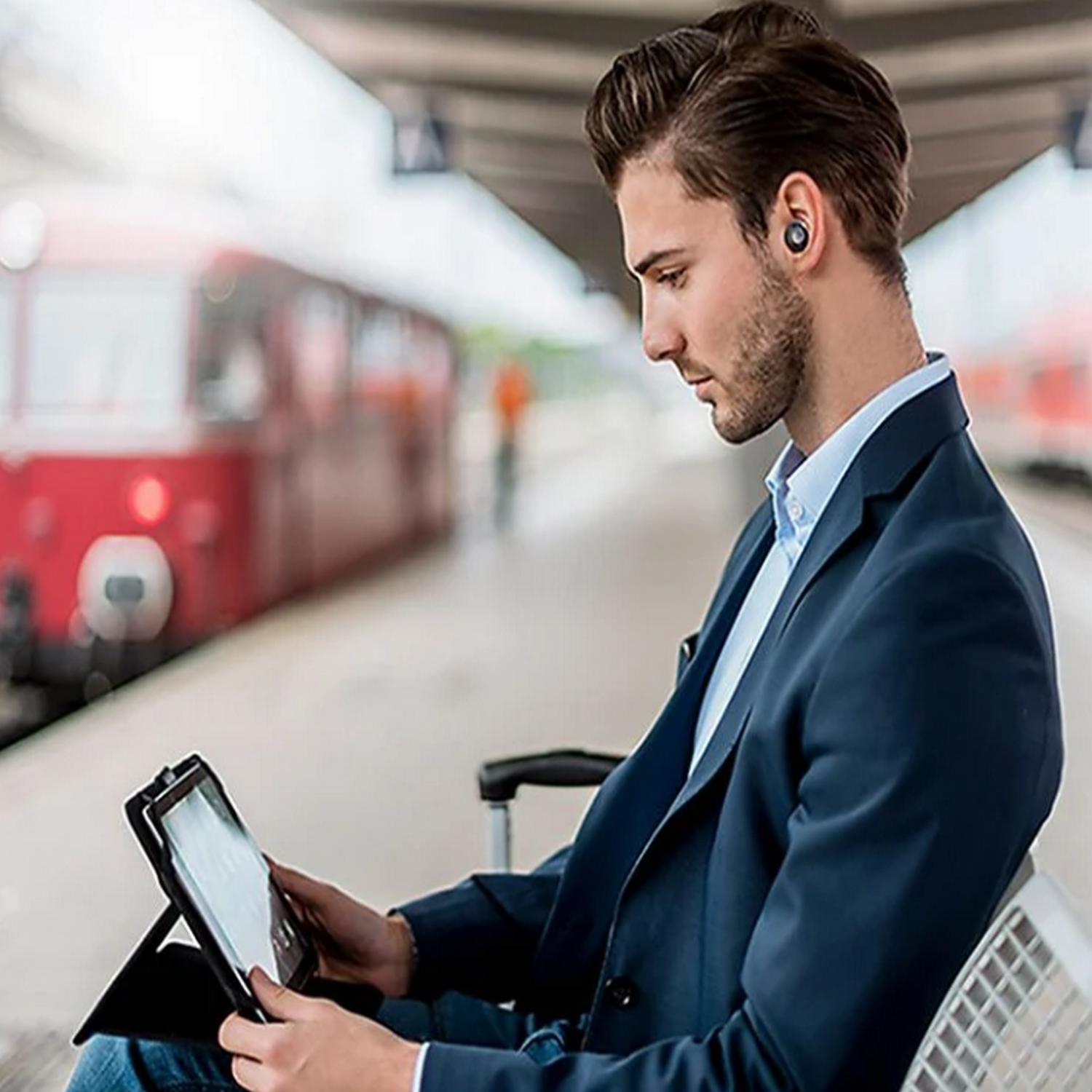 Man met oortjes, kijkend naar een tablet, zittend in een treinstation.