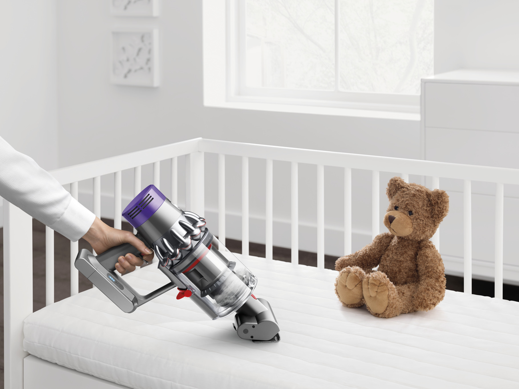 Persona aspirando el colchón de un bebé con una aspiradora Dyson, junto a un oso de peluche.