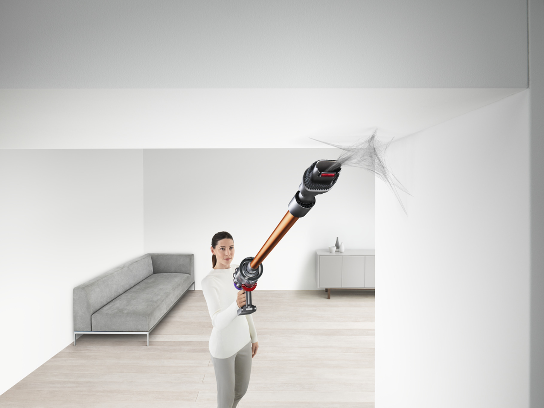 Mujer aspira el techo con Dyson. Sofá gris y pared blanca al fondo.
