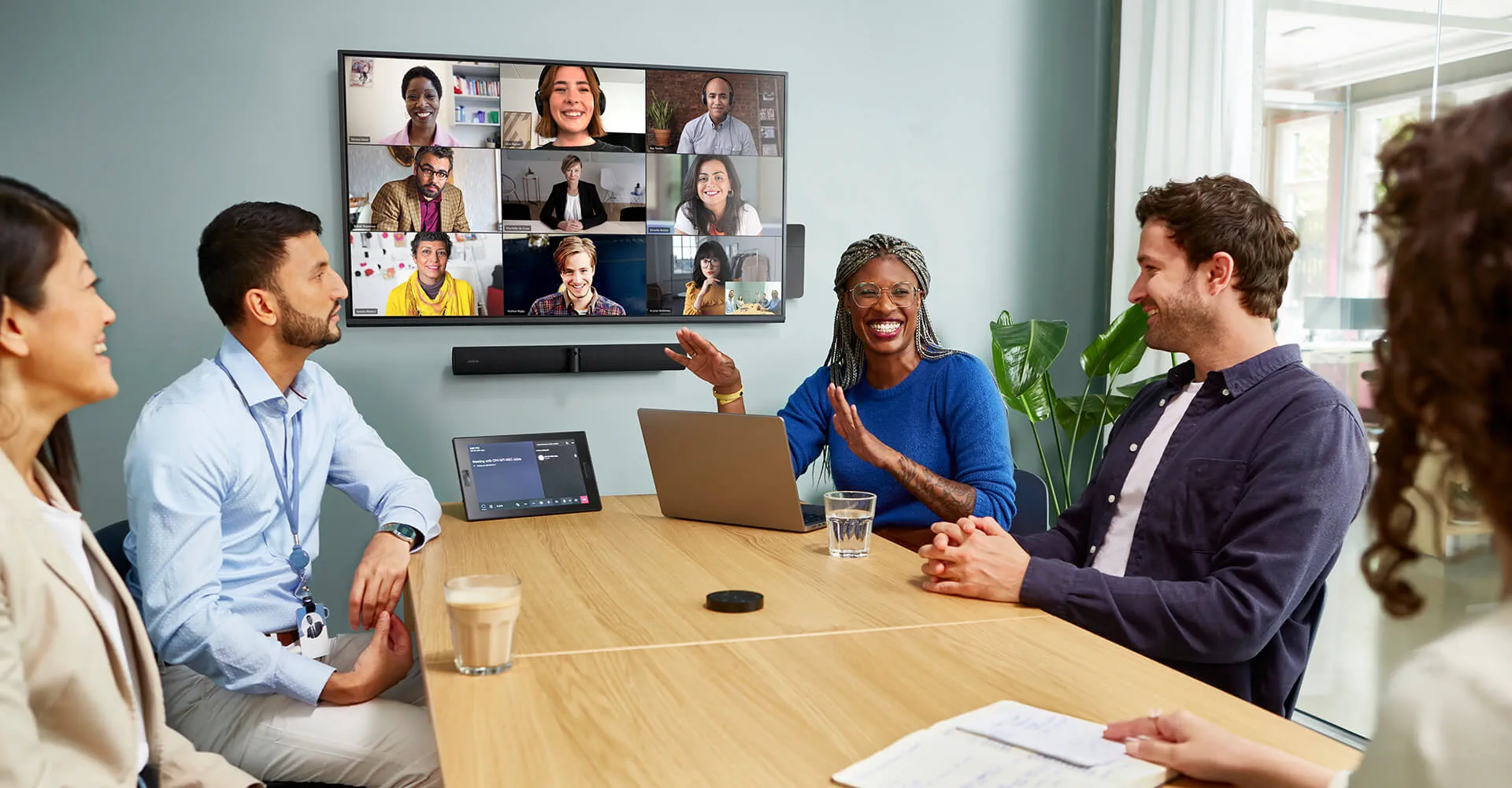 Ein Treffen von Personen in einem Videoanruf auf einem großen Bildschirm. Personen sitzen um einen Tisch.