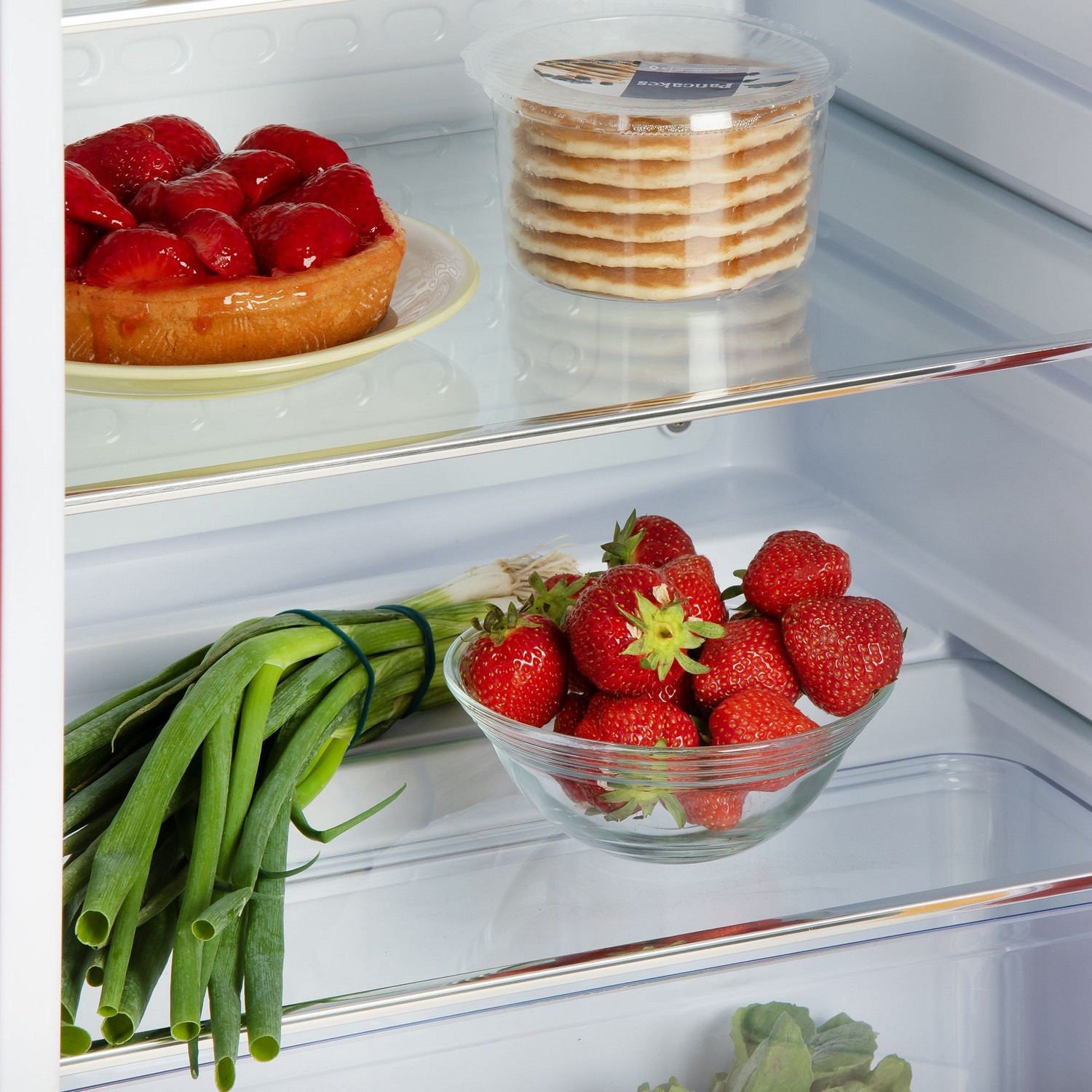 Close up of foods in a refrigerator: strawberries, a tart, and a container of pancakes.