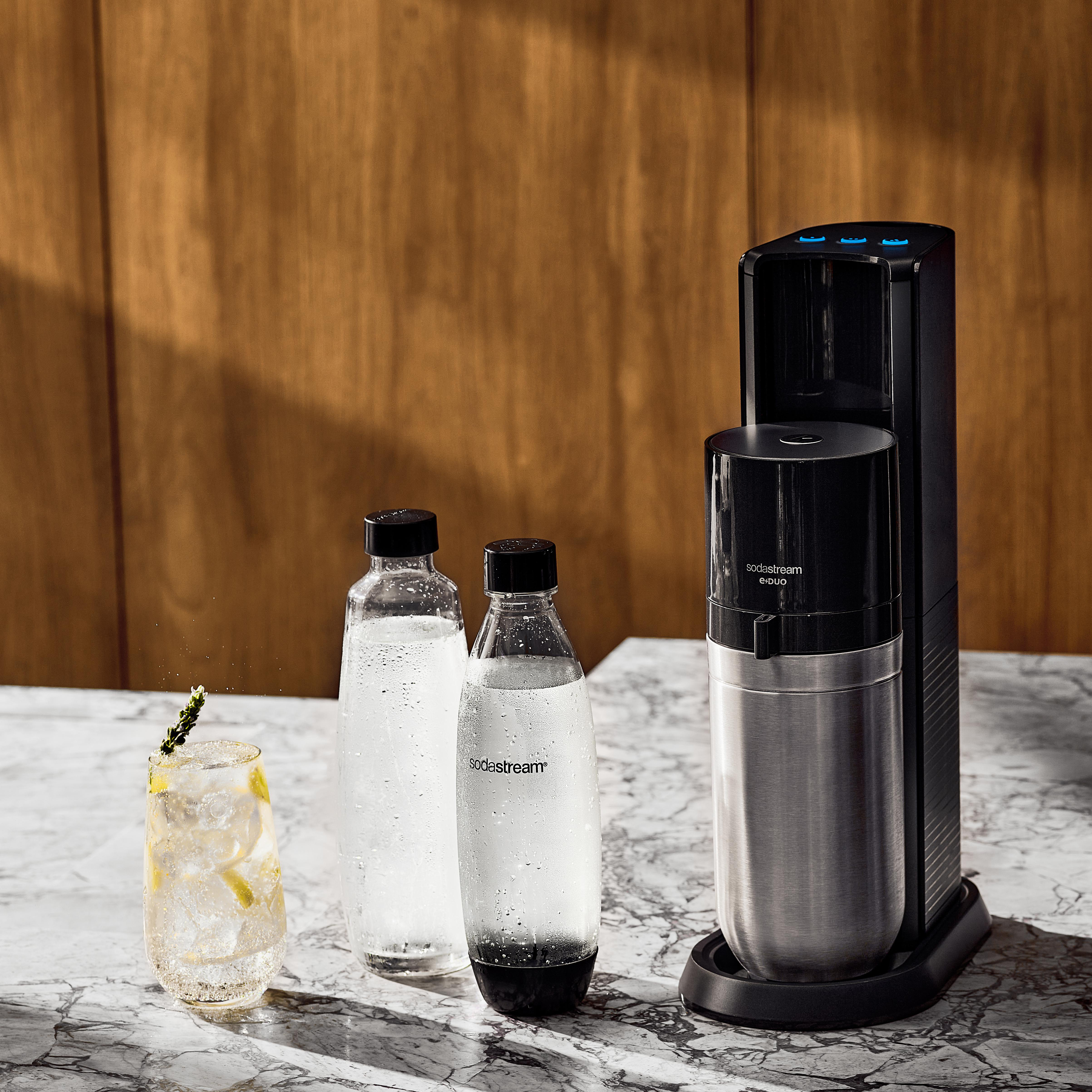 SodaStream-Maschine mit Wasserflaschen und einem Glas Getränk auf Marmoroberfläche.