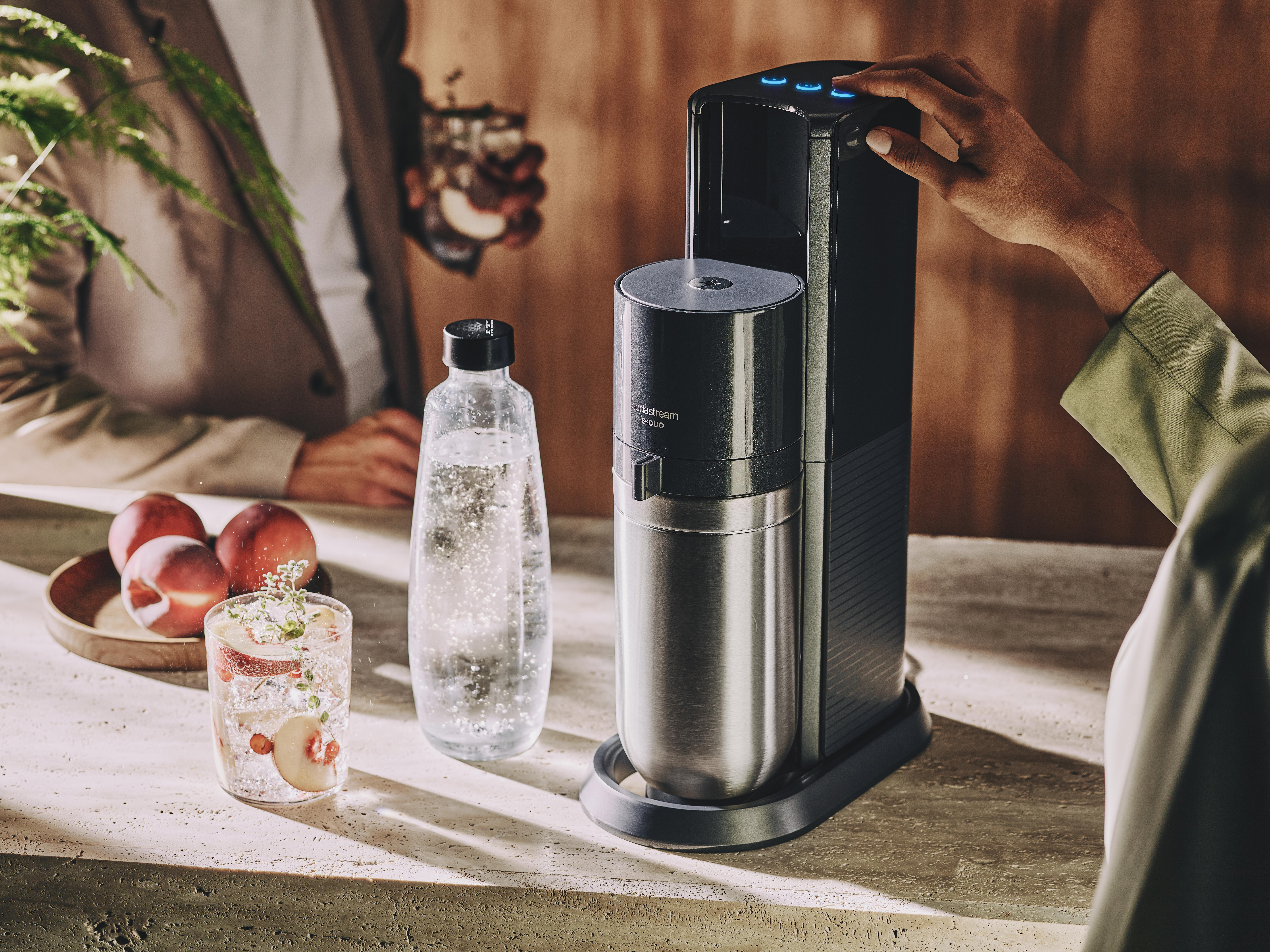 Person benutzt SodaStream, mit einem Glas Getränk, Wasserflasche und Pfirsichen auf einem Tisch.