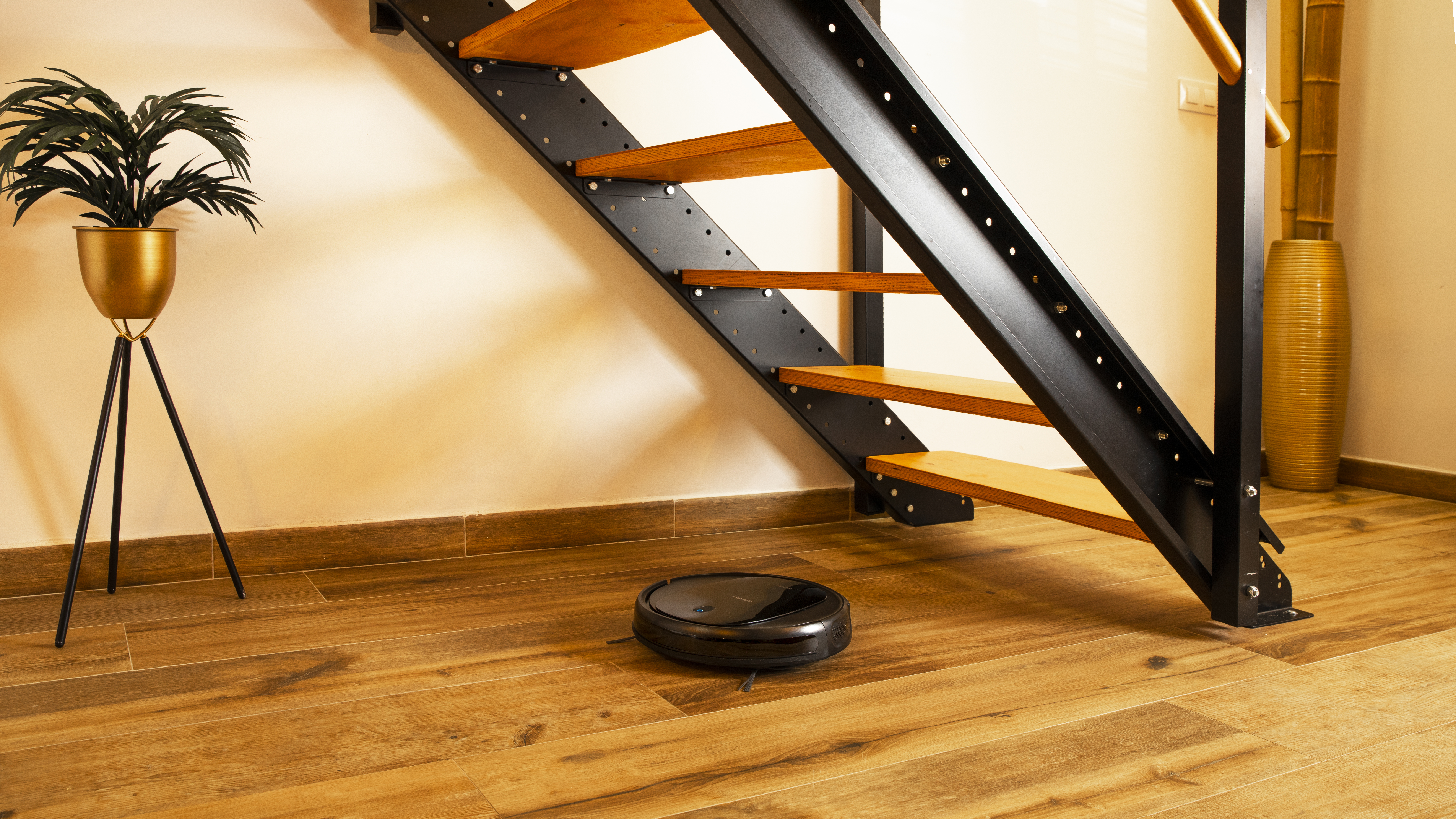 Aspiradora robot en suelo de madera cerca de escaleras con una planta y jarrón de bambú.