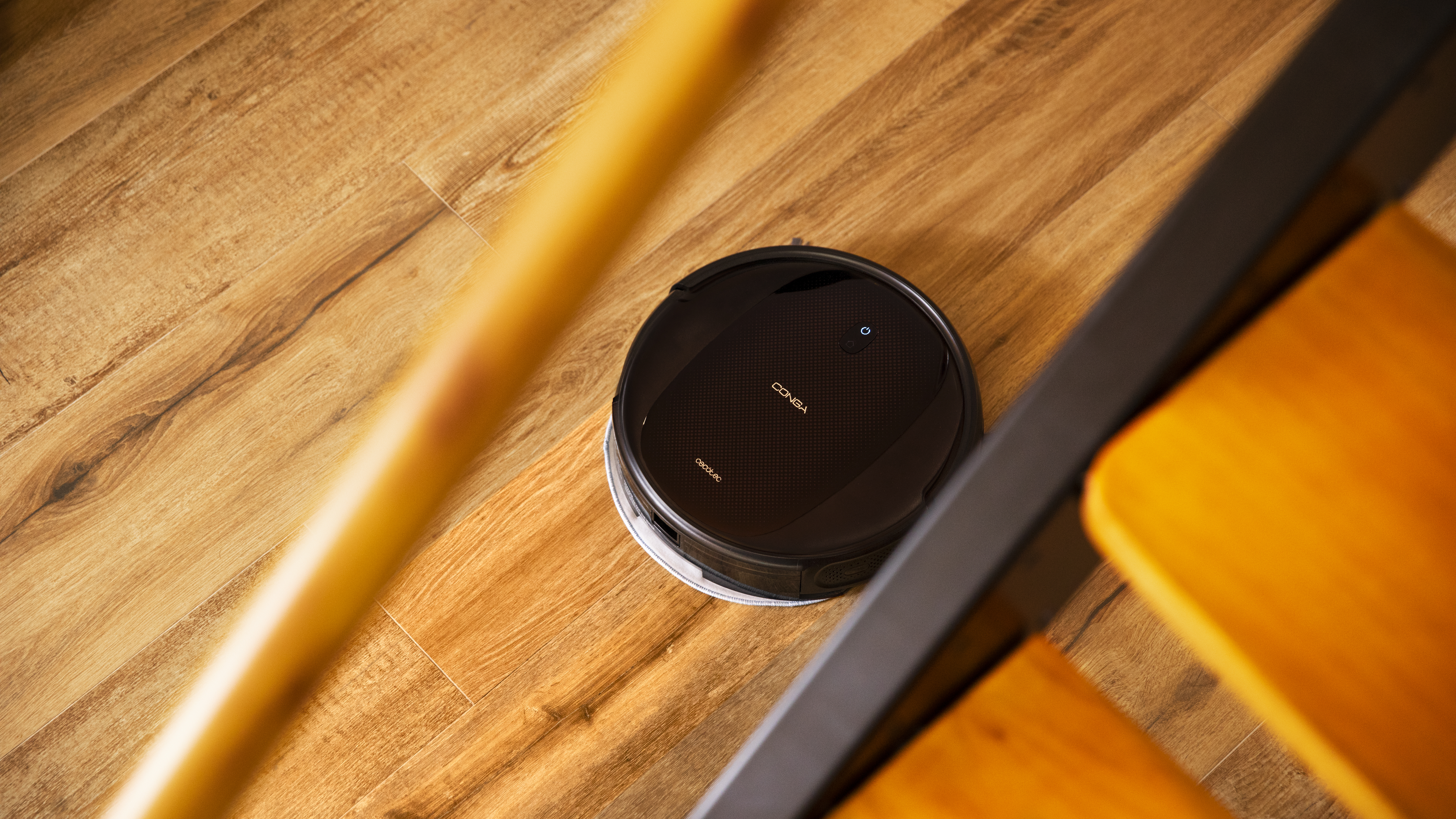 Aspiradora robot negra en suelo de madera. Pata de silla amarillo-naranja en el marco.