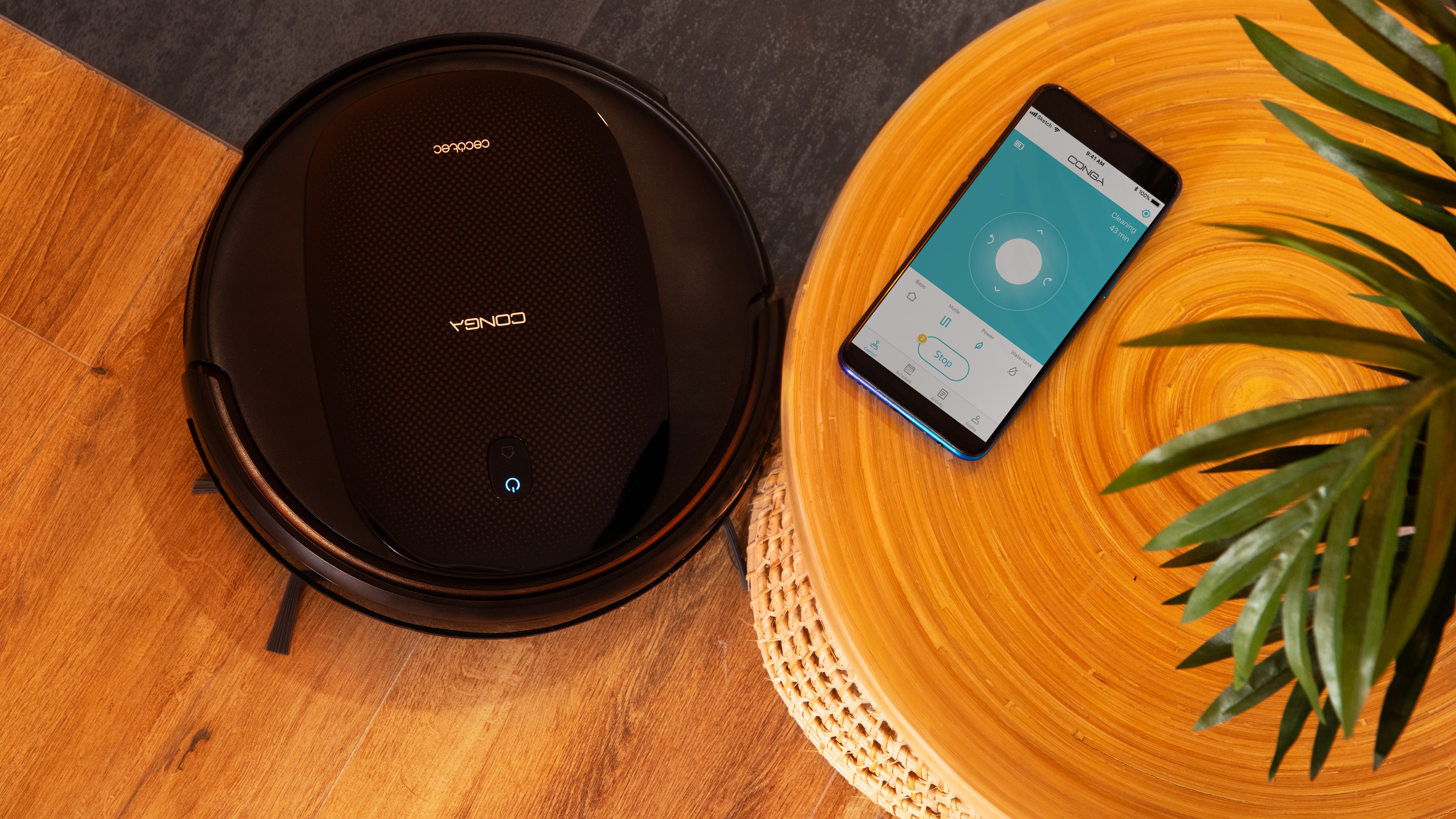 Aspiradora robot negra sobre suelo de madera, con teléfono mostrando la aplicación.