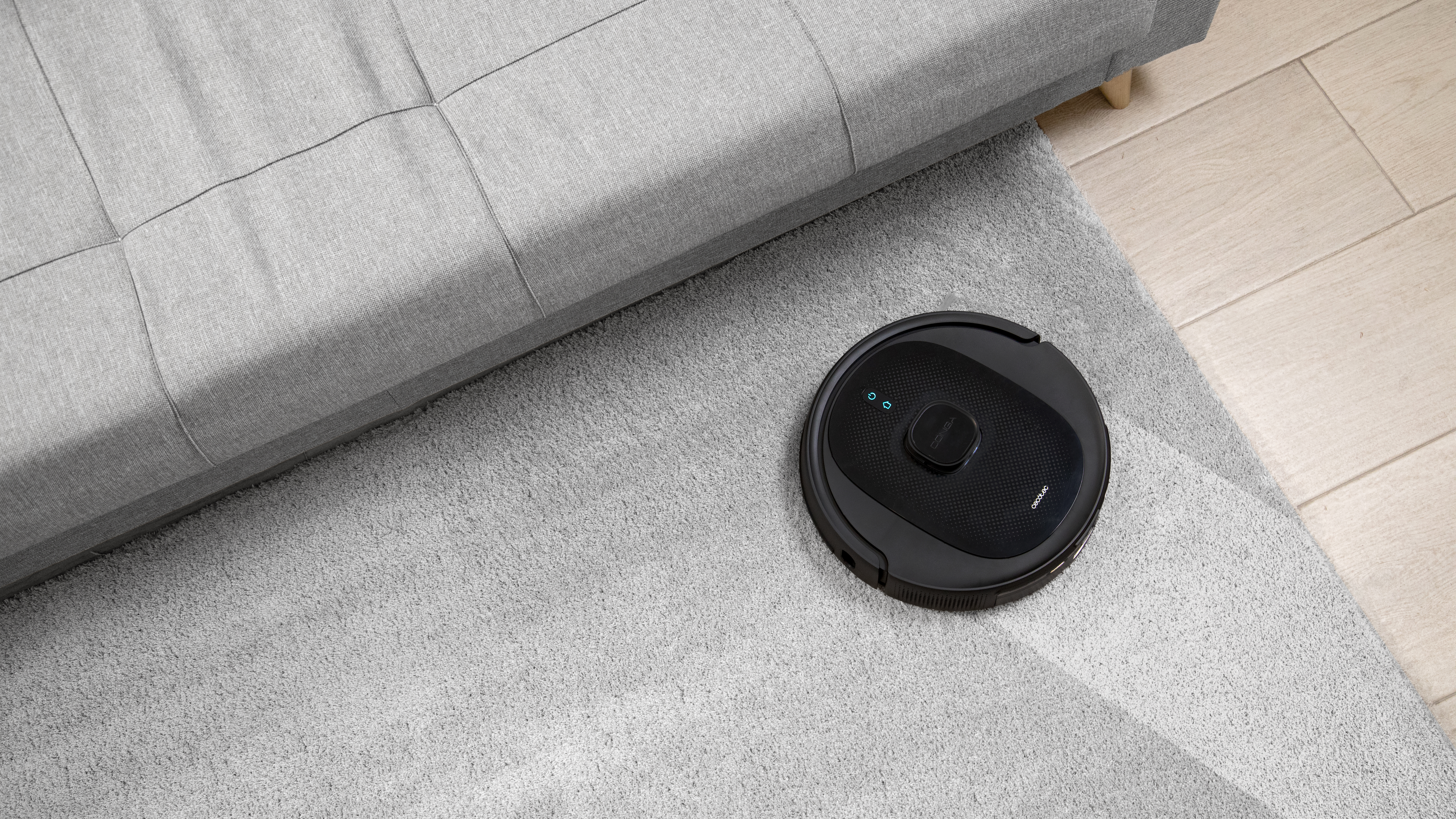 Una aspiradora robot negra sobre una alfombra junto a un sofá.