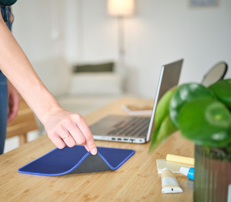Egy kéz egy kék egérpadot tart egy faasztalon, laptop, növény és művészeti kellékek mellett.