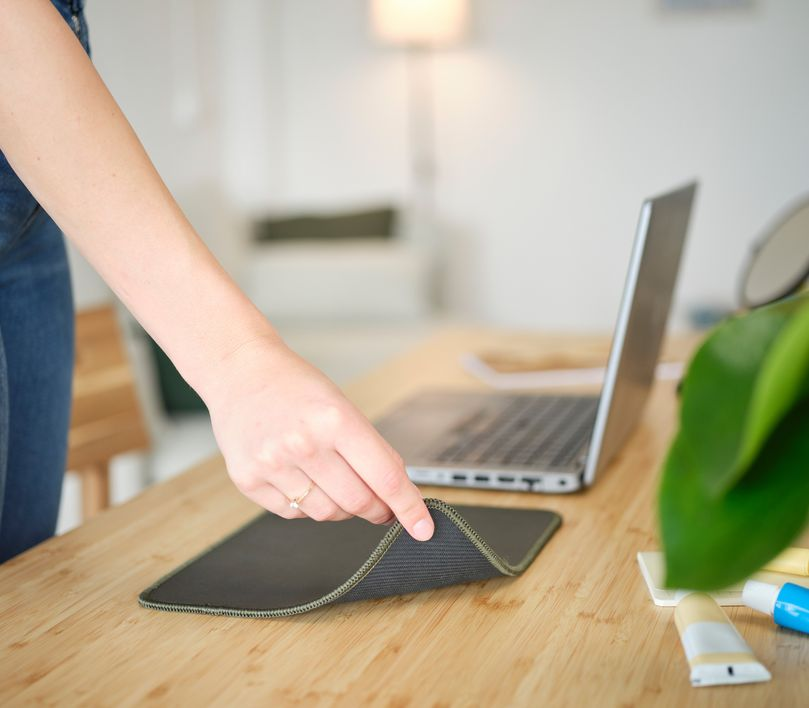 Egy kéz tart egy fekete egérpadot egy fa asztalon, egy laptop, és növények közelében.