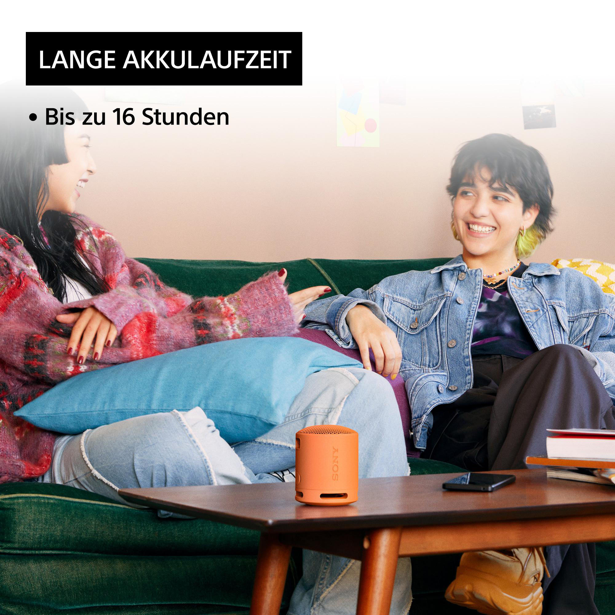 Zwei Frauen auf einem Sofa mit einem orangefarbenen Lautsprecher, Text: Lange Akkulaufzeit.