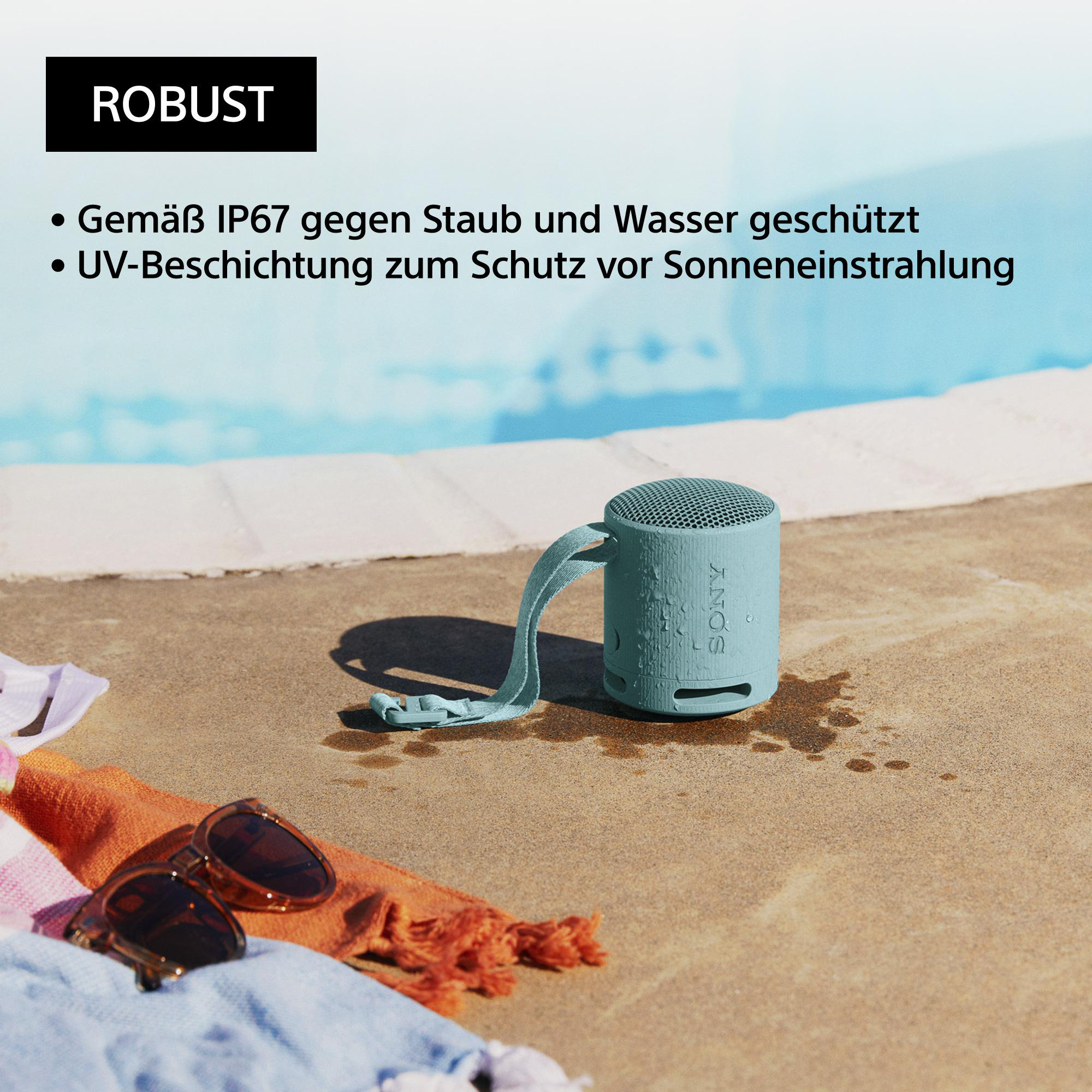 Ein türkisfarbener tragbarer Lautsprecher mit Griff, auf einer Betonoberfläche in der Nähe eines Pools, mit Sonnenbrille und Handtuch.