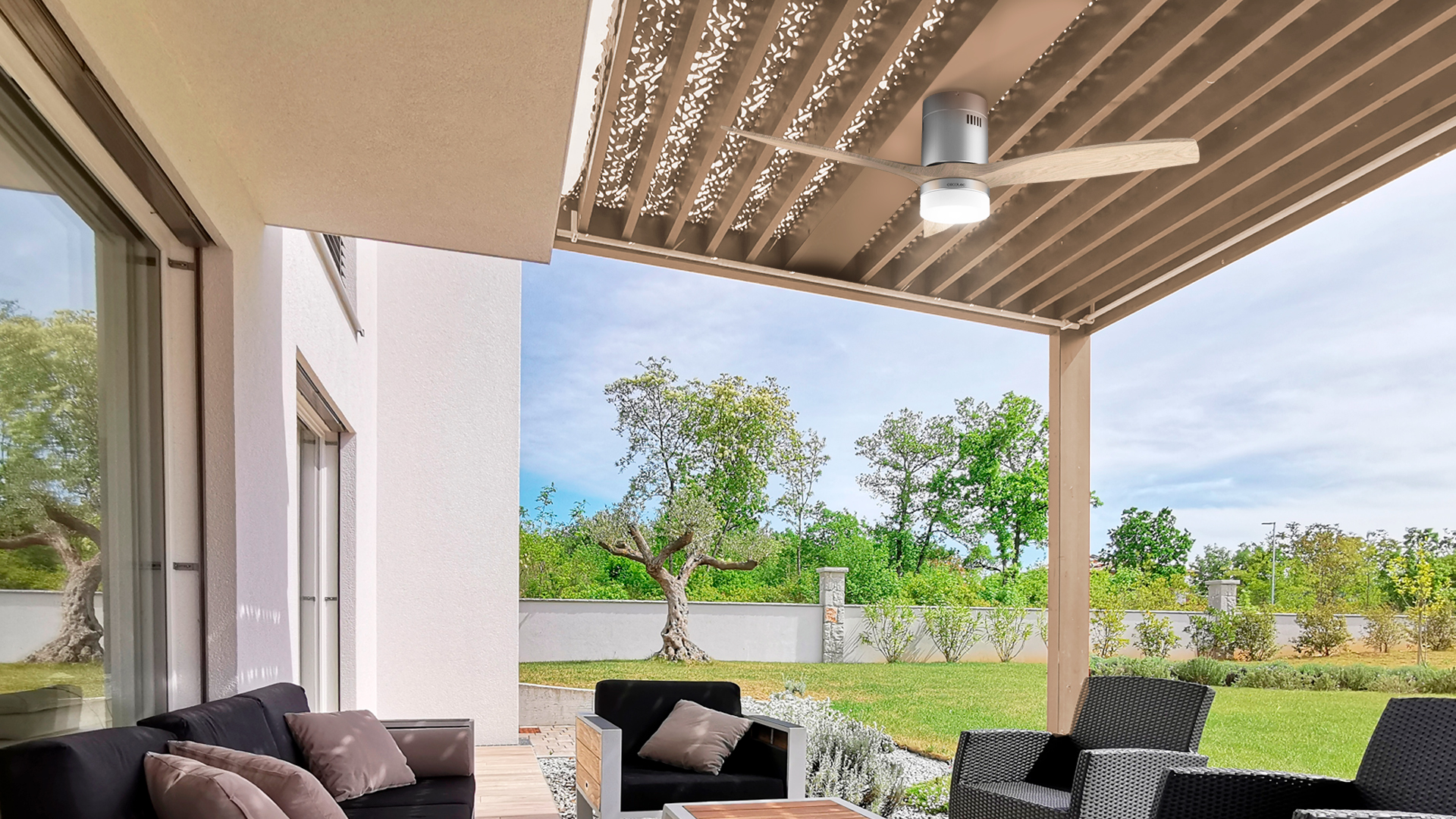 Un patio al aire libre con ventilador, asientos y vista a un jardín con árboles y cielo.