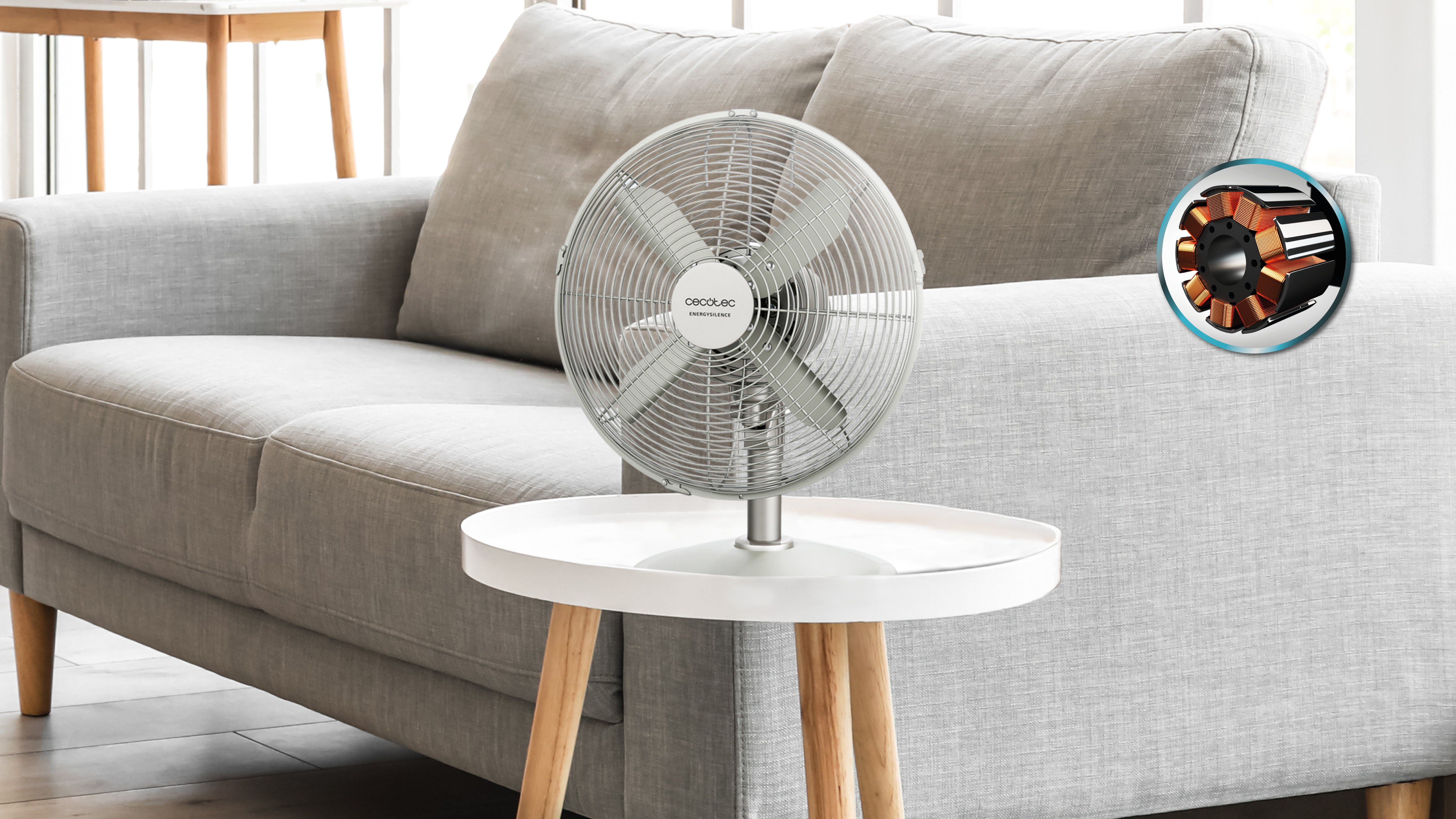 Un ventilador plateado sobre una mesa blanca. Un sofá gris en el fondo. Primer plano del motor.