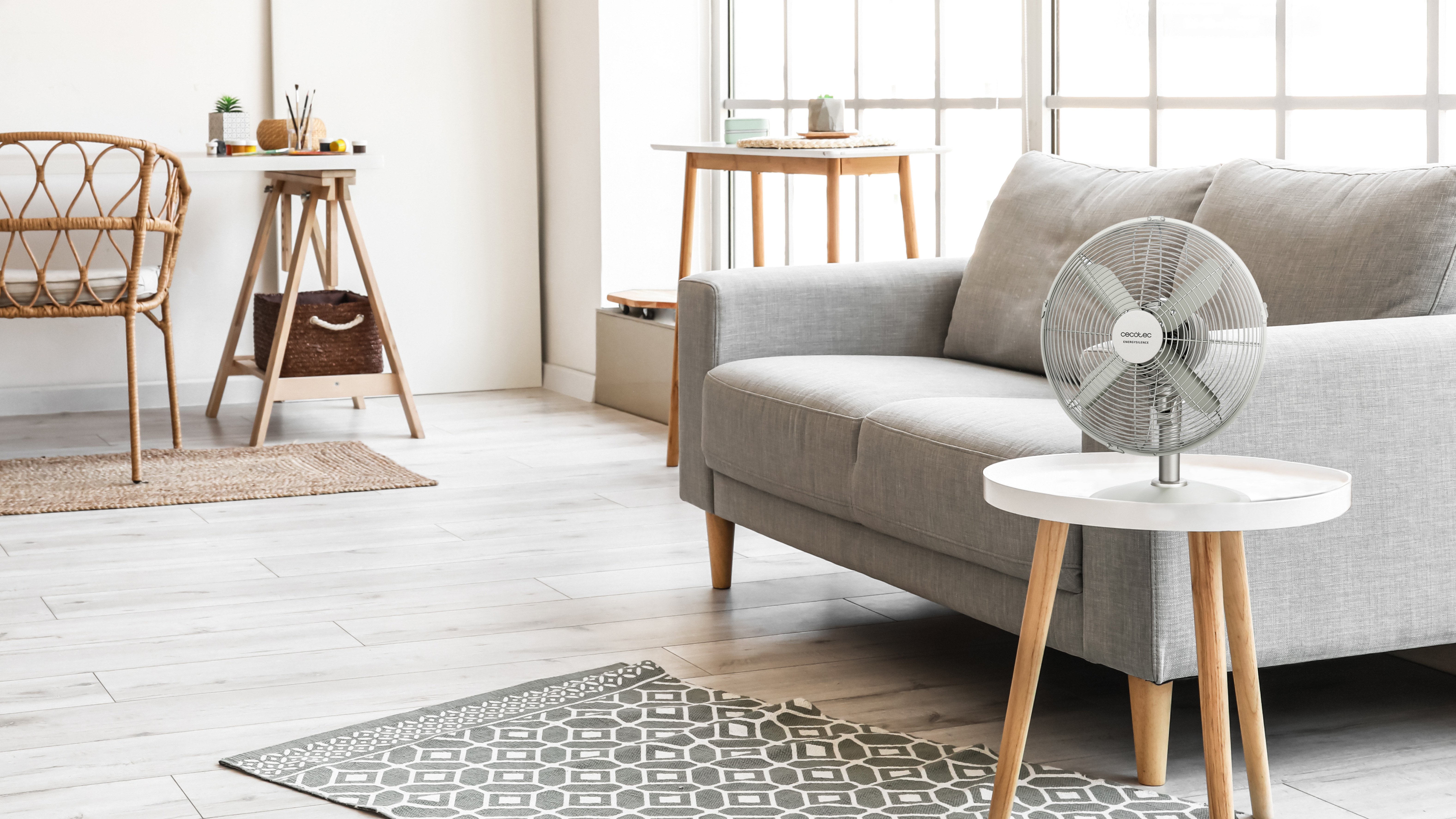 Interior de habitación con sofá gris, ventilador en mesa, alfombra y ventana.
