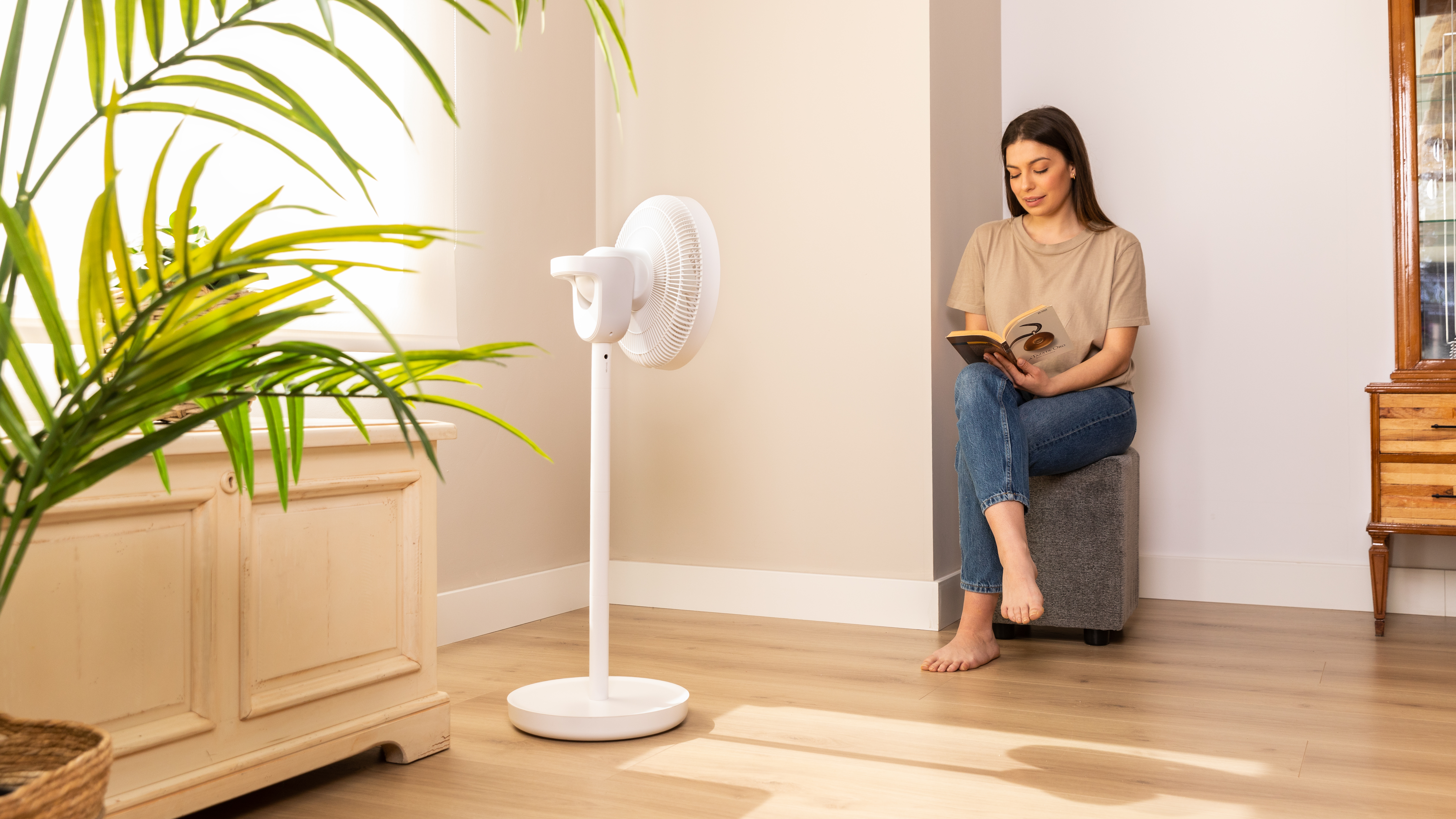 Una mujer lee un libro junto a un ventilador blanco. Una planta verde en el fondo.