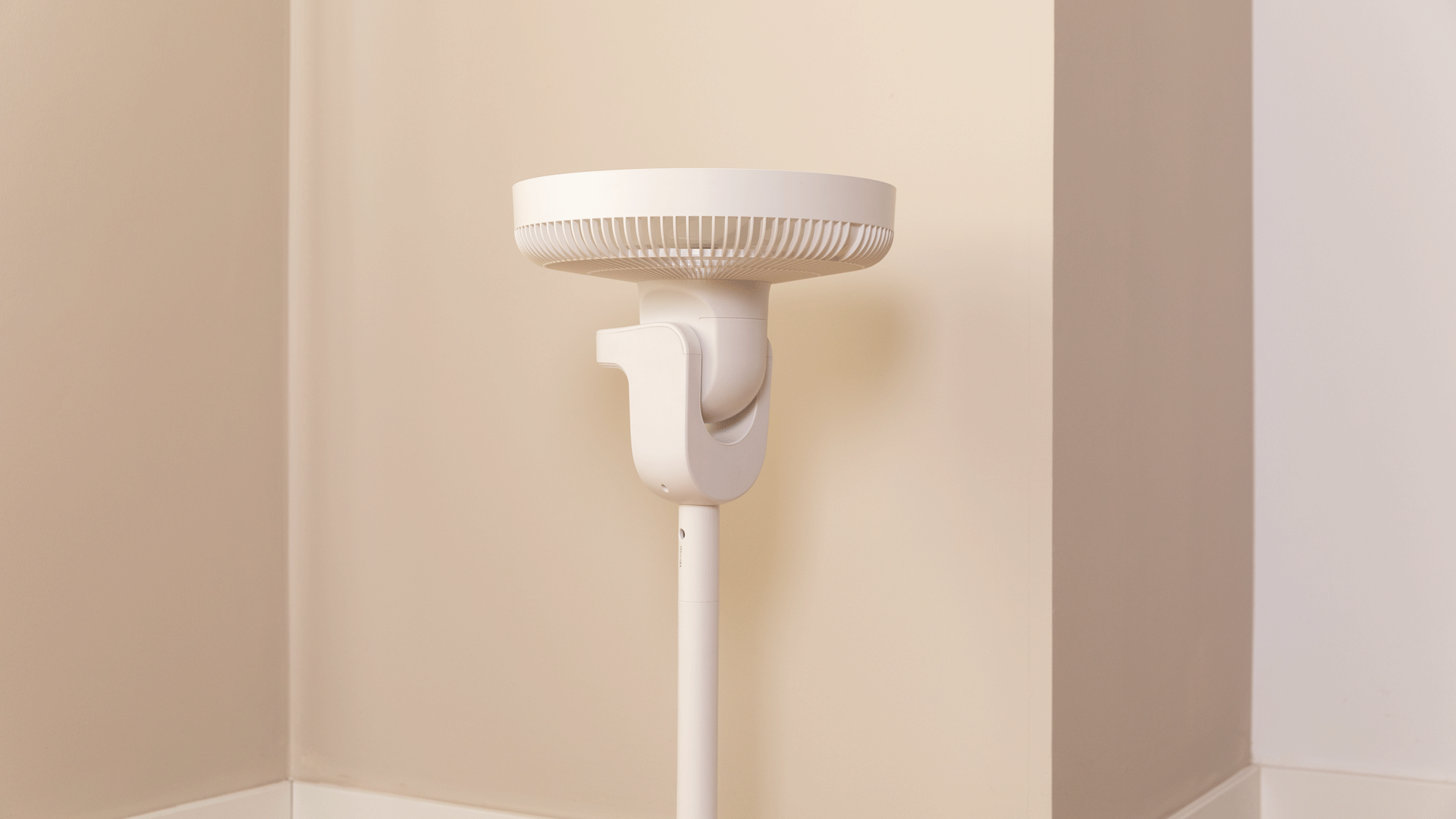 Ventilador blanco sobre un soporte en la esquina de una habitación, contra una pared beige.