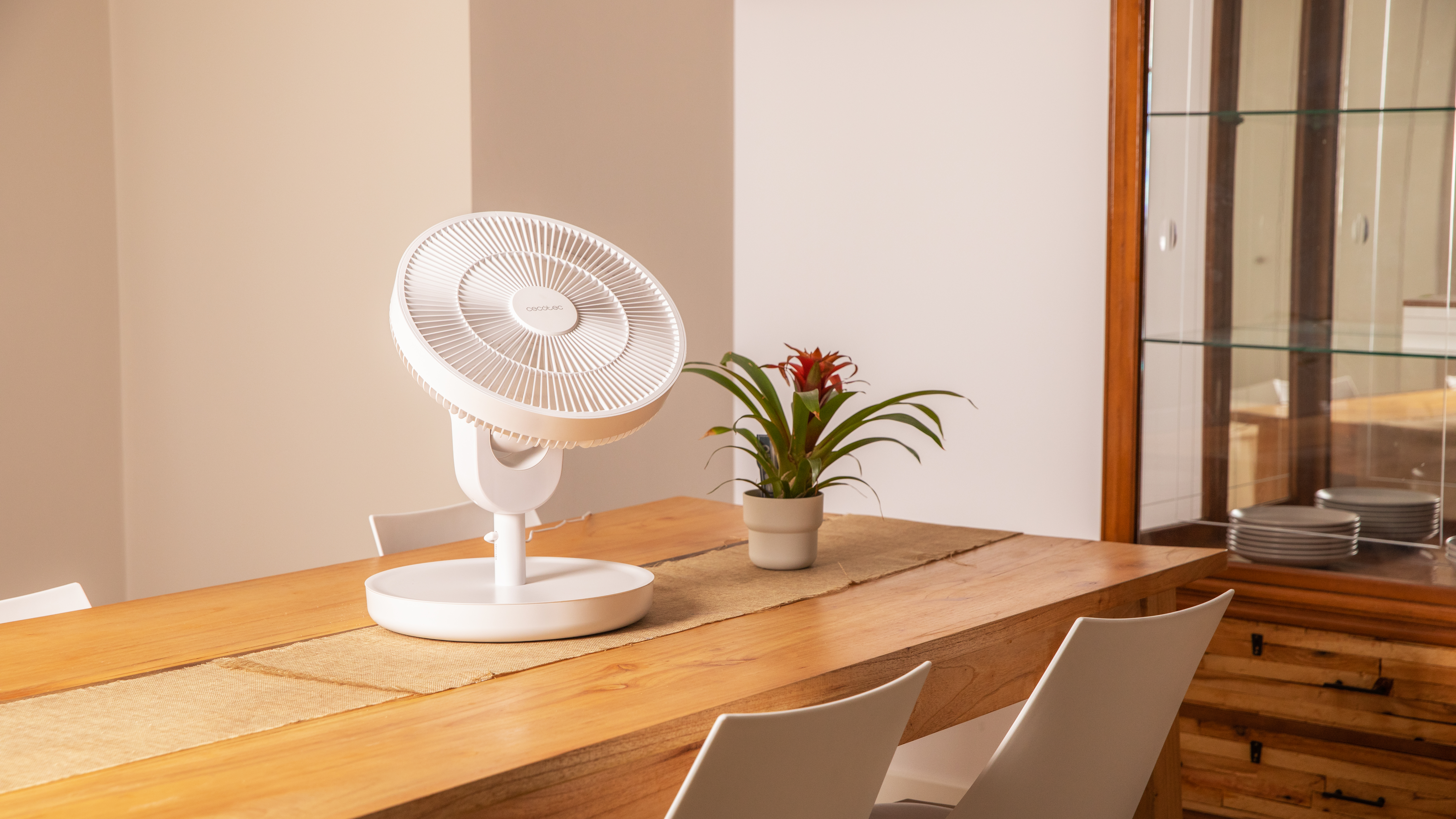 Ventilador blanco sobre una mesa de madera. Una planta y platos en la mesa, sillas alrededor.