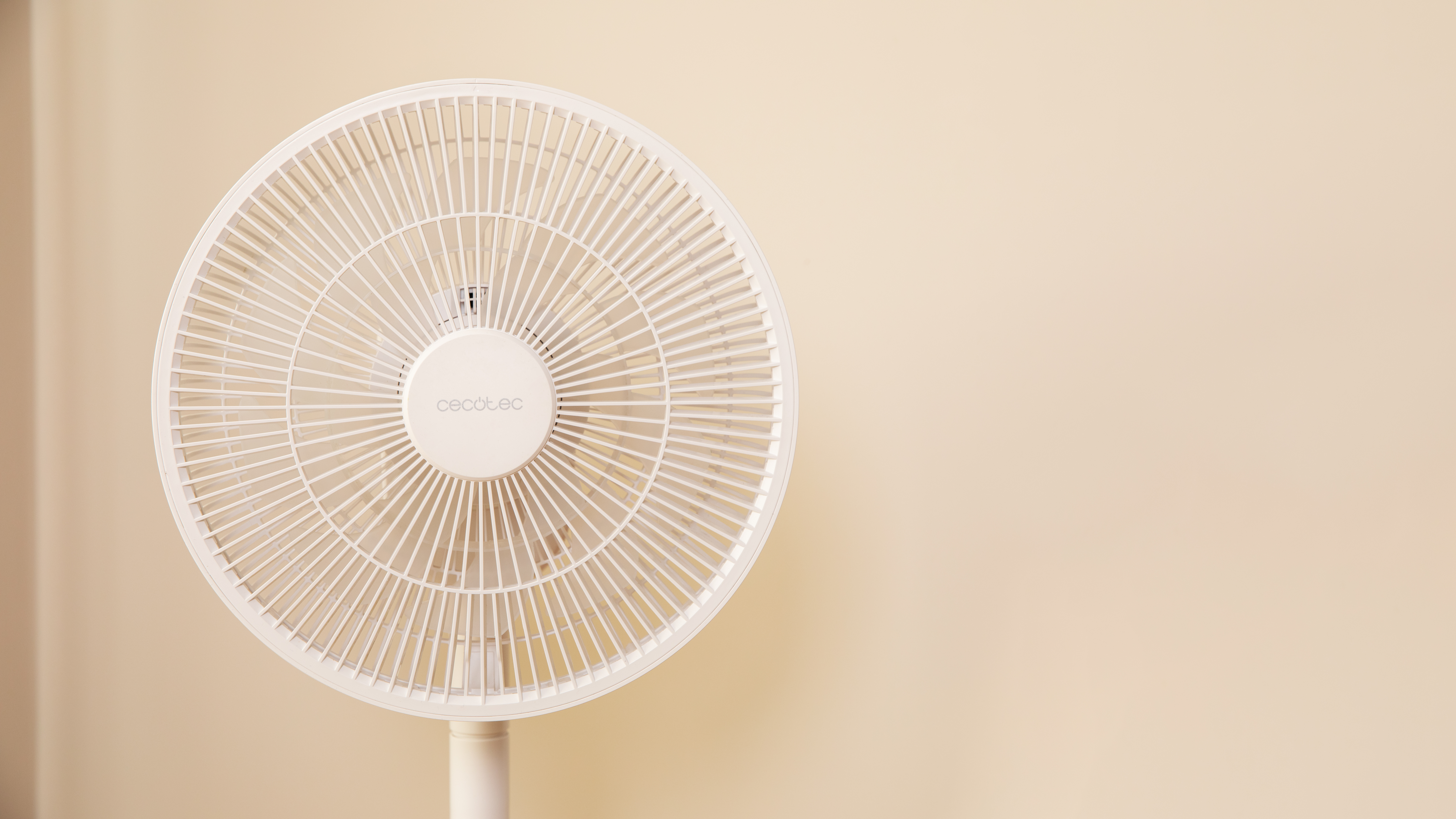 Un ventilador de pie blanco con cabezal circular, contra una pared beige claro.