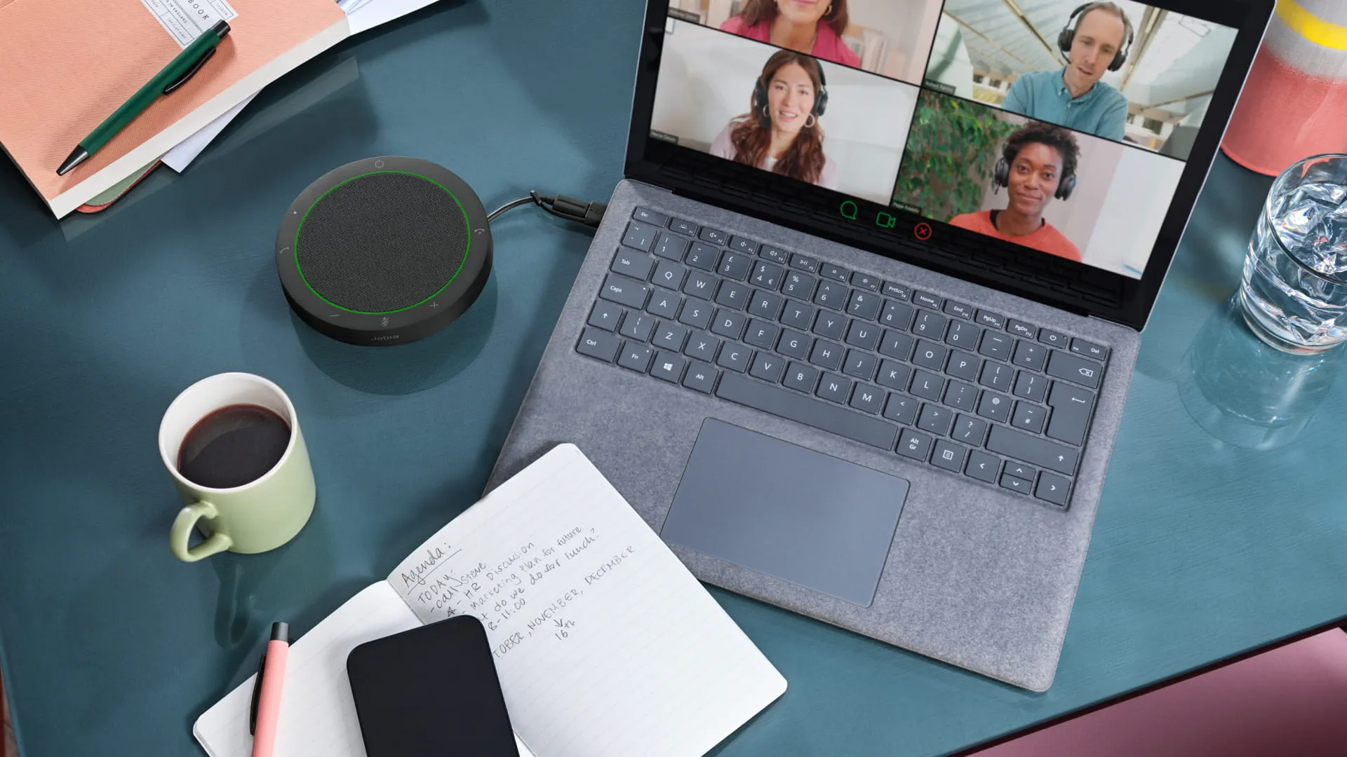Laptop zeigt einen Videoanruf mit 5 Personen, Lautsprecher, Notizblock, Kaffee und Wasserglas.