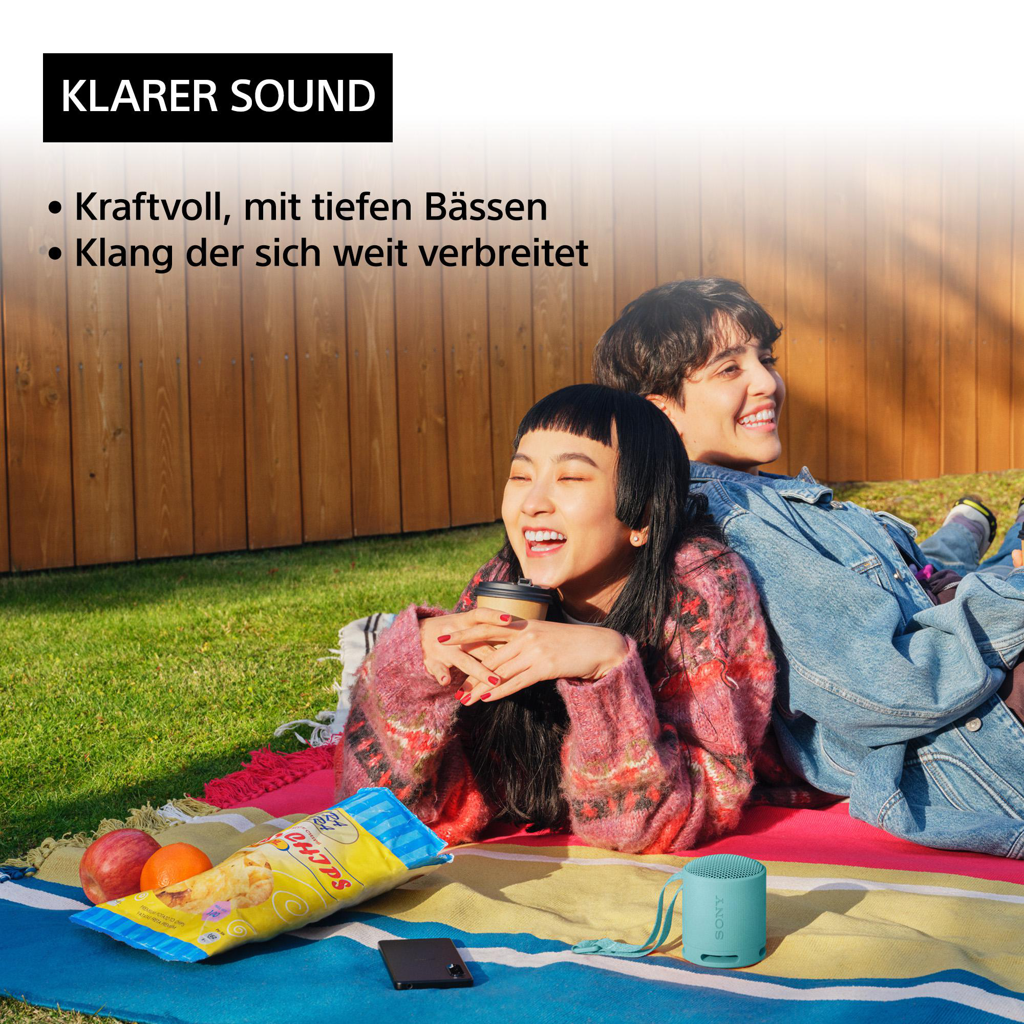 Zwei lächelnde Personen mit blauem Lautsprecher, Snack und Obst auf bunter Decke draußen.