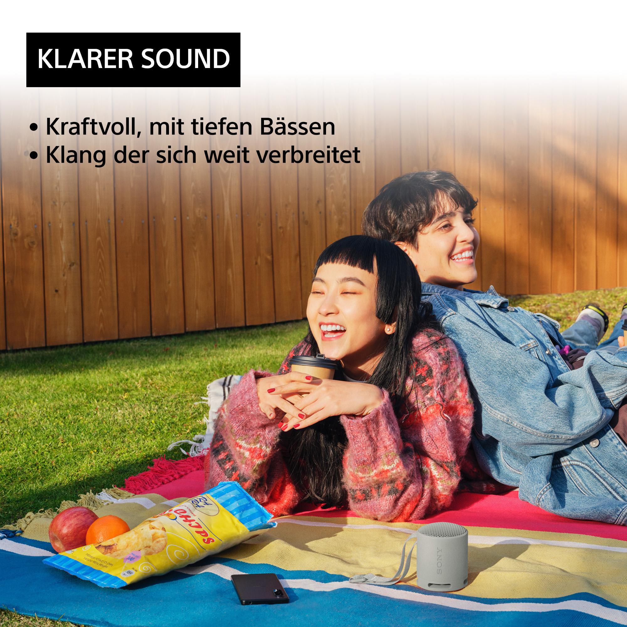 Zwei Personen lachen auf einer Picknickdecke mit Snacks und einem Lautsprecher.